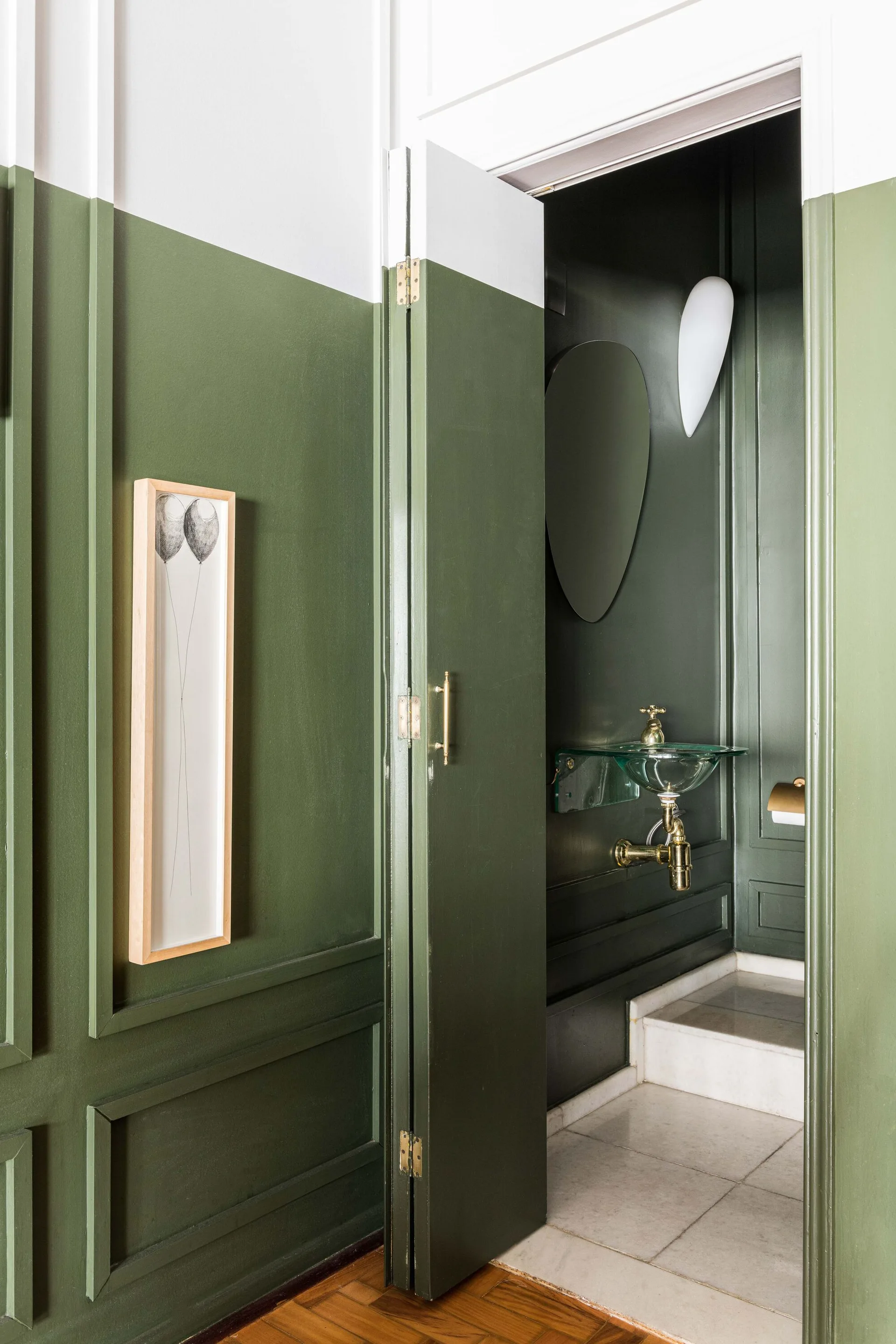 An open green paneled door reveals a small powder room with a glass sink, brass fixtures, and a white sconce.