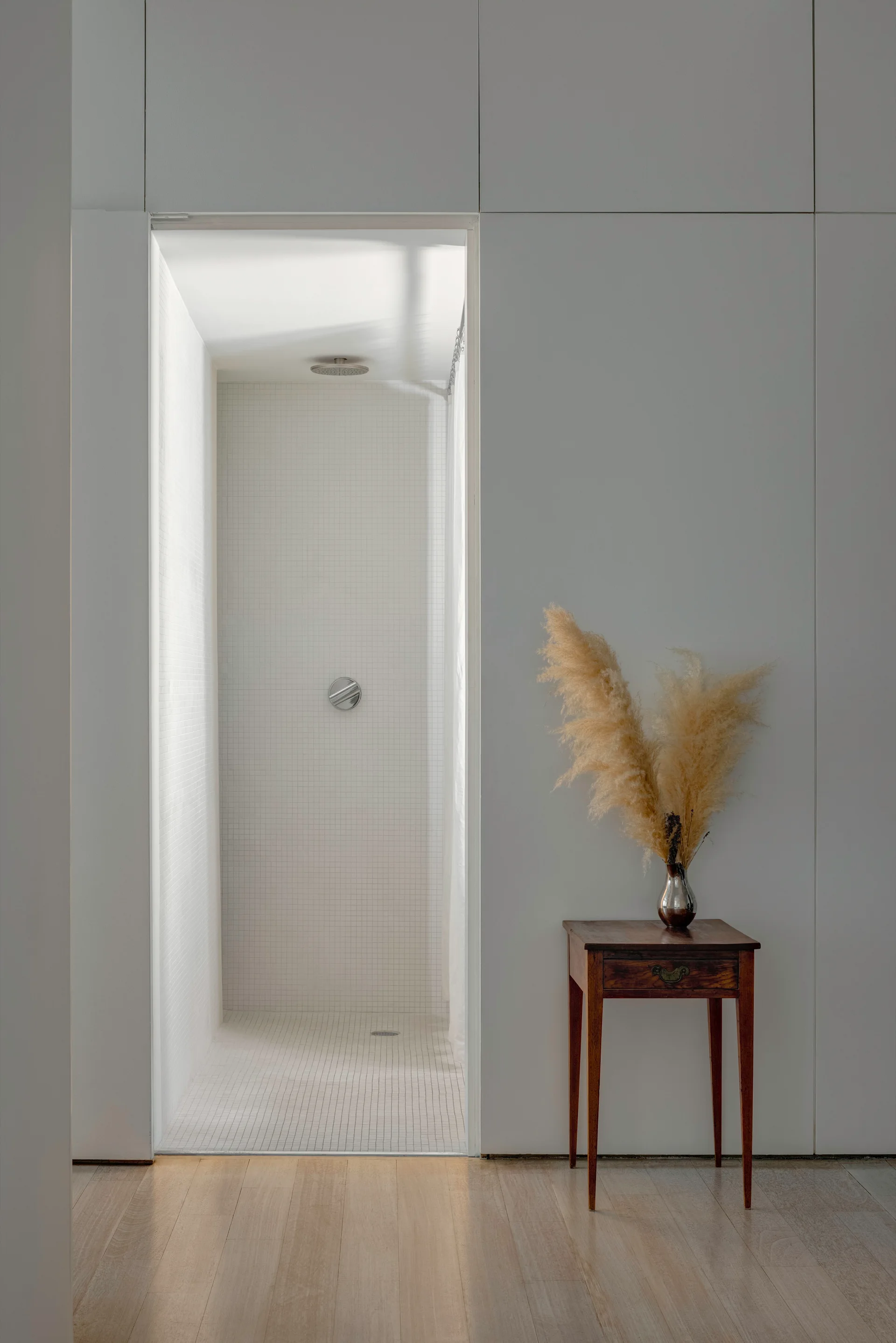 A minimalist bathroom features a recessed shower stall with white mosaic tiles next to a dark wood console table holding pampas grass.