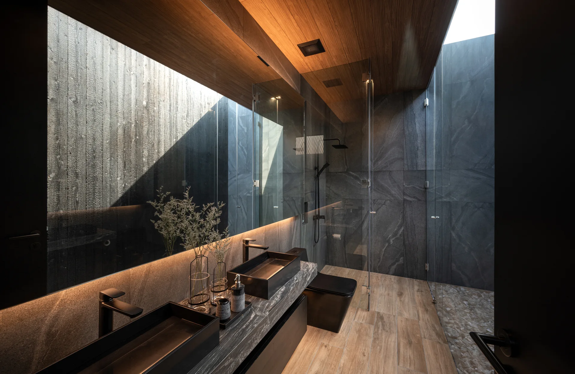 A modern dark bathroom with dual black vessel sinks on a stone vanity, a backlit mirror, wood flooring, and a glass shower under a skylight.