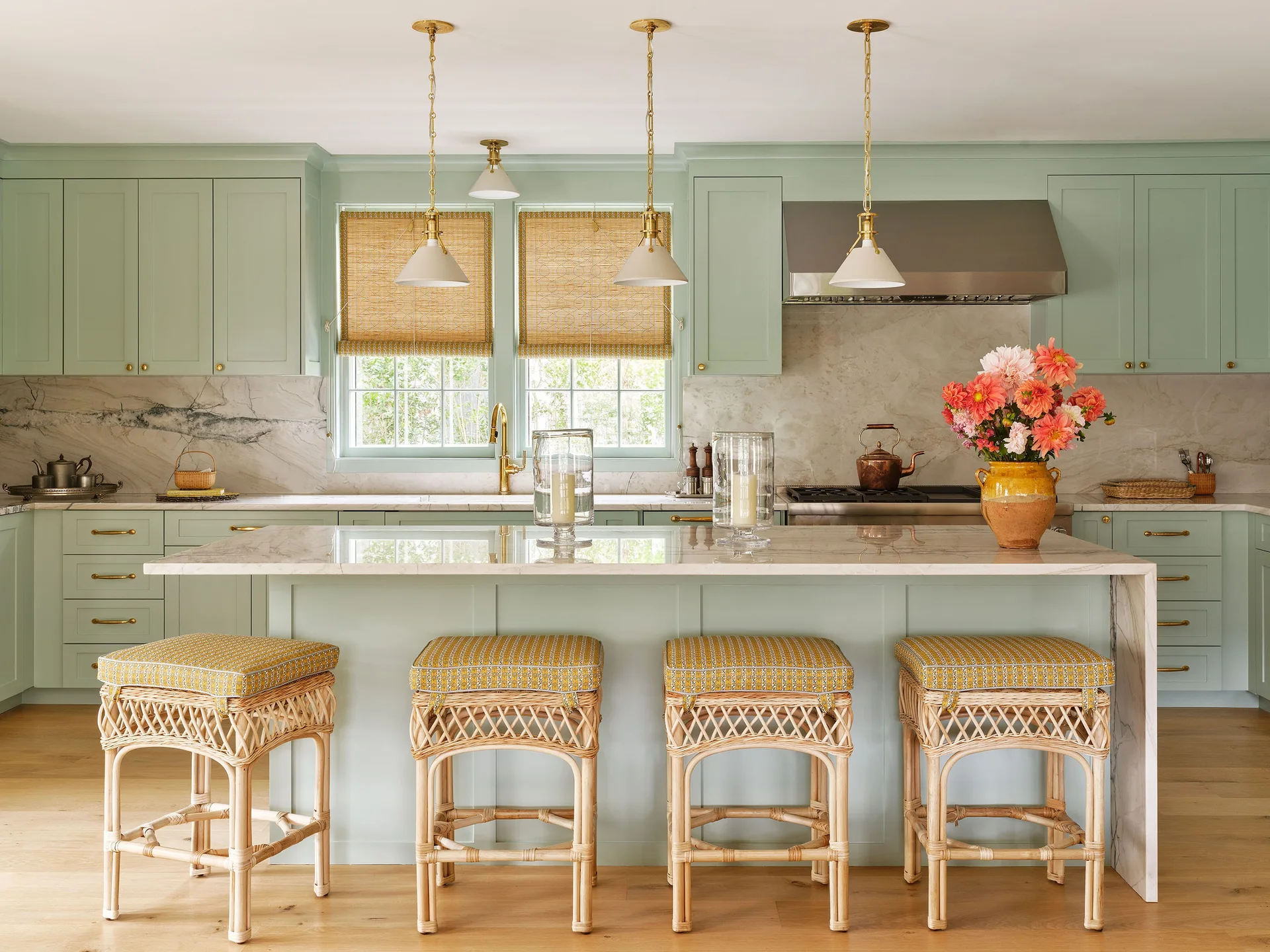 Mint green kitchen cabinetry surrounds a marble-topped island paired with four rattan bar stools under brass pendant lights.