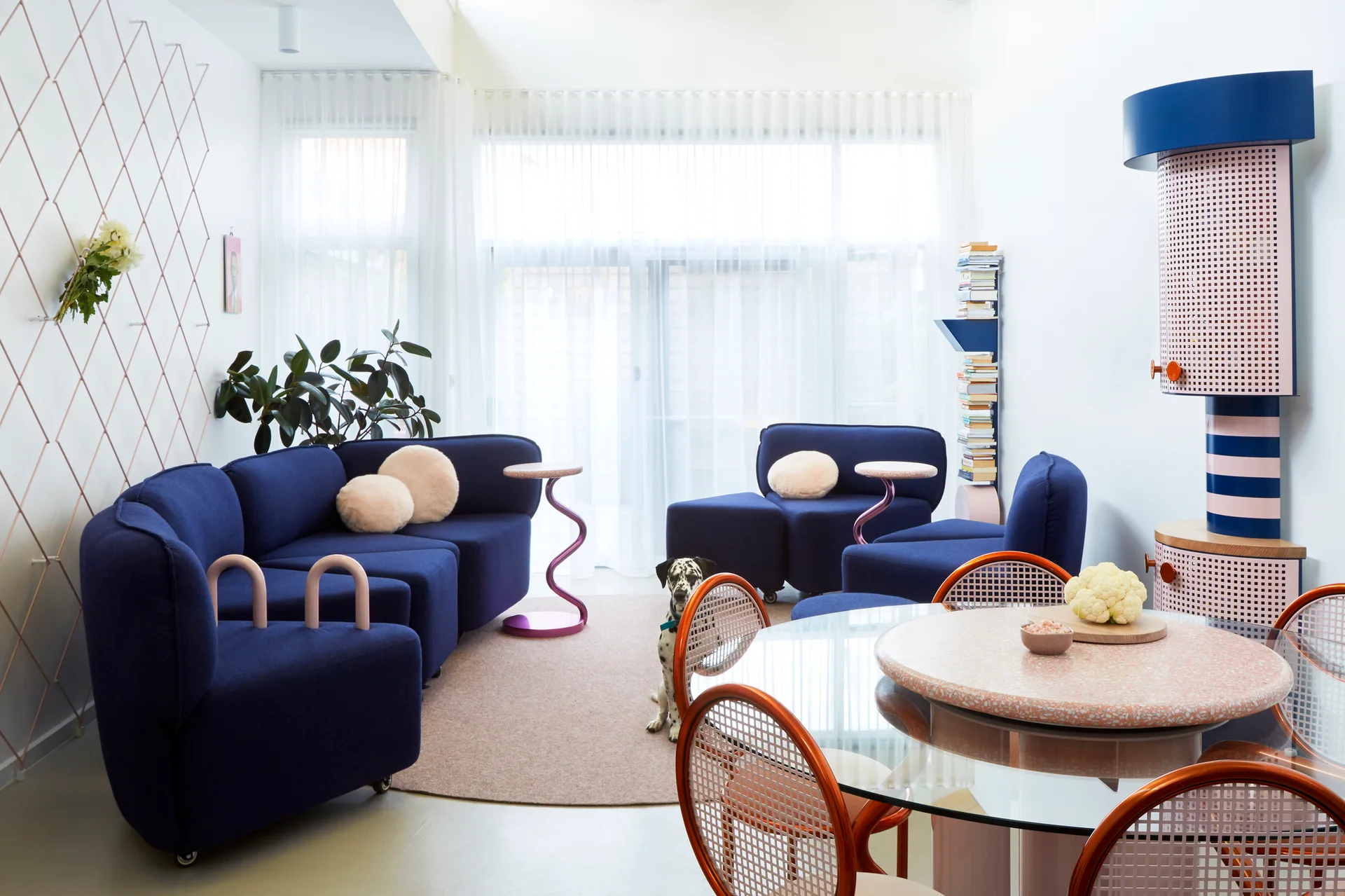 A bright living room features blue modular seating, pink wavy side tables, and a glass-topped dining table set against sheer white curtains.