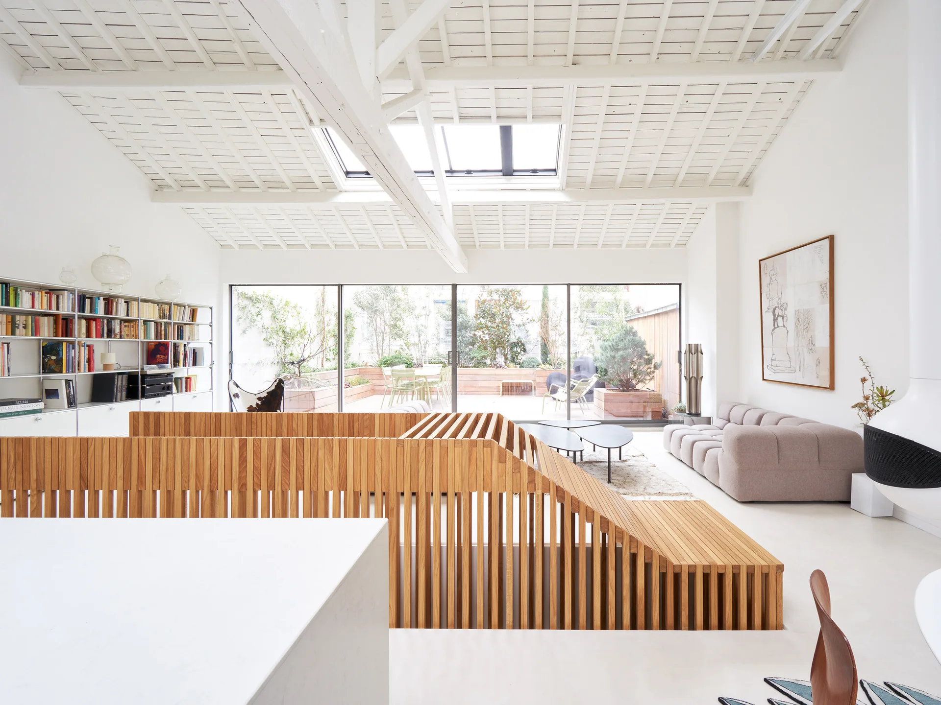 A bright white living room with a slatted wood staircase railing, a modular bookshelf, a beige sofa, and a skylight in the sloped ceiling.