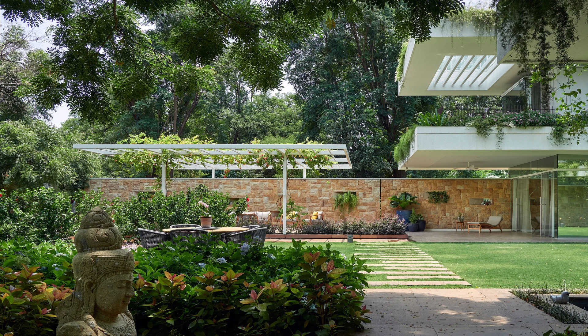 A landscaped backyard garden features a lush green lawn with a stepping stone path, a white pergola over a patio, and a textured tan stone wall.