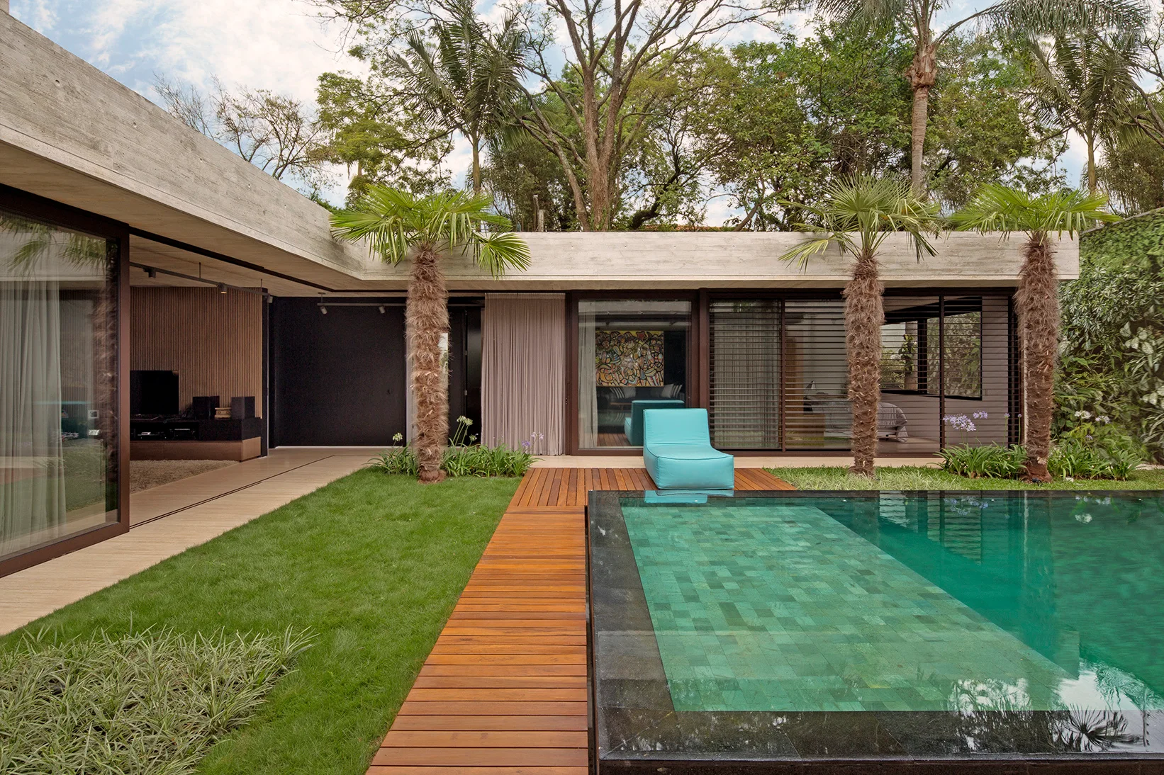 A modern courtyard features a rectangular swimming pool with teal tiles, wooden plank decking, a green lawn, and palm trees under a concrete roof.