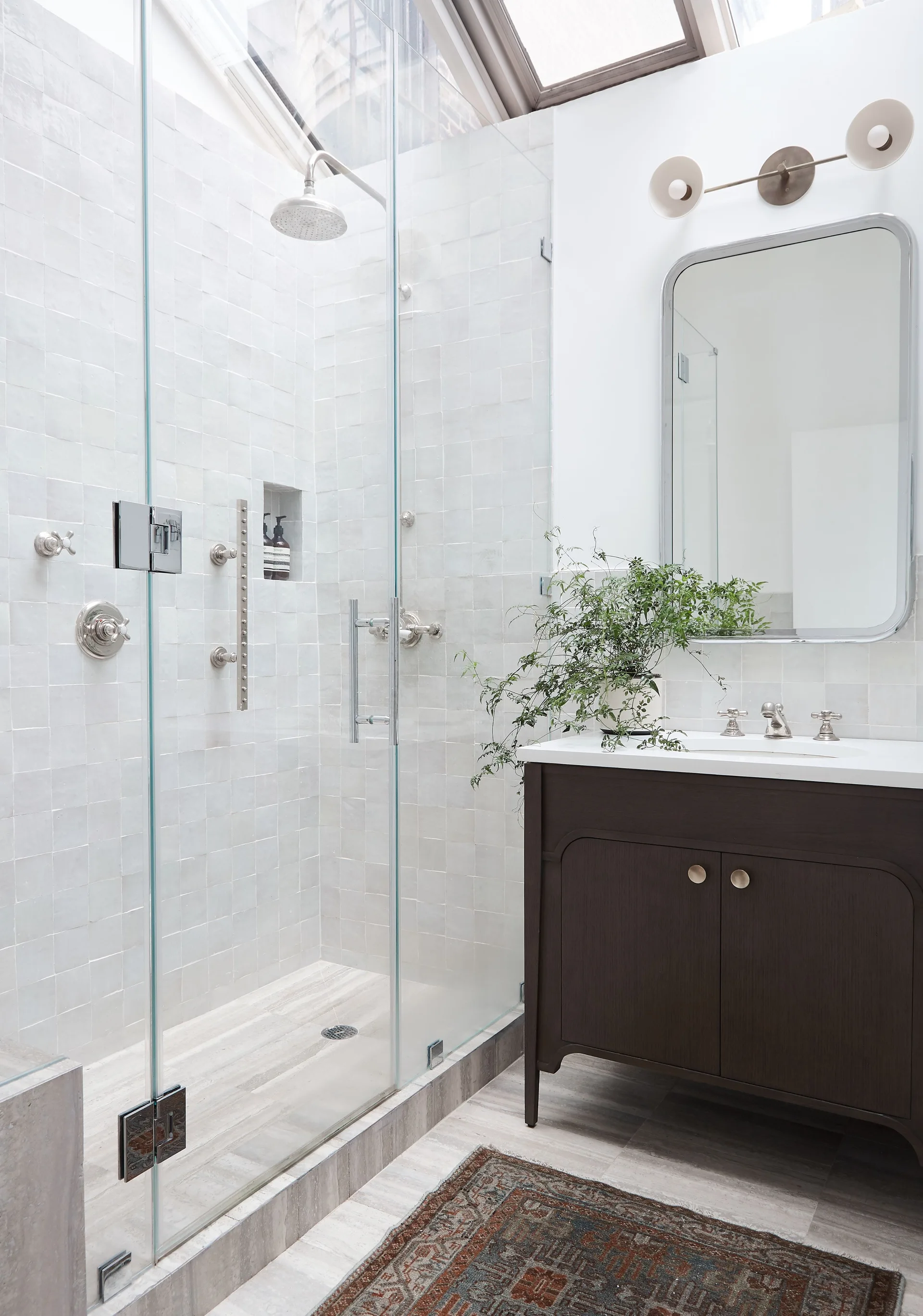 A modern bathroom features a dark wood vanity with a rounded mirror, a glass shower enclosure with square white tiles, and a skylight overhead.