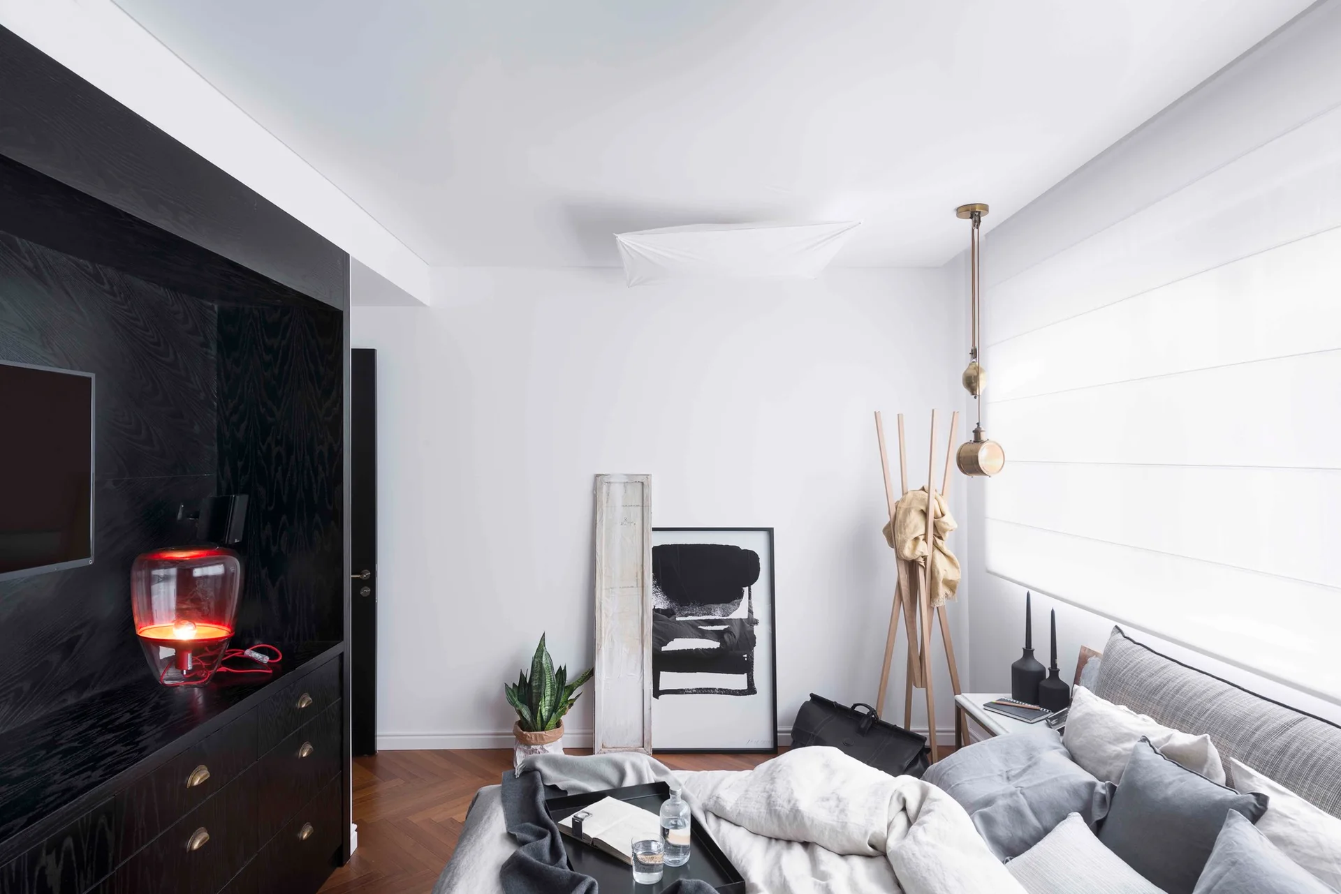 A modern bedroom features white bedding, a wooden herringbone floor, and a large black wooden built-in cabinet with a red glass lamp.