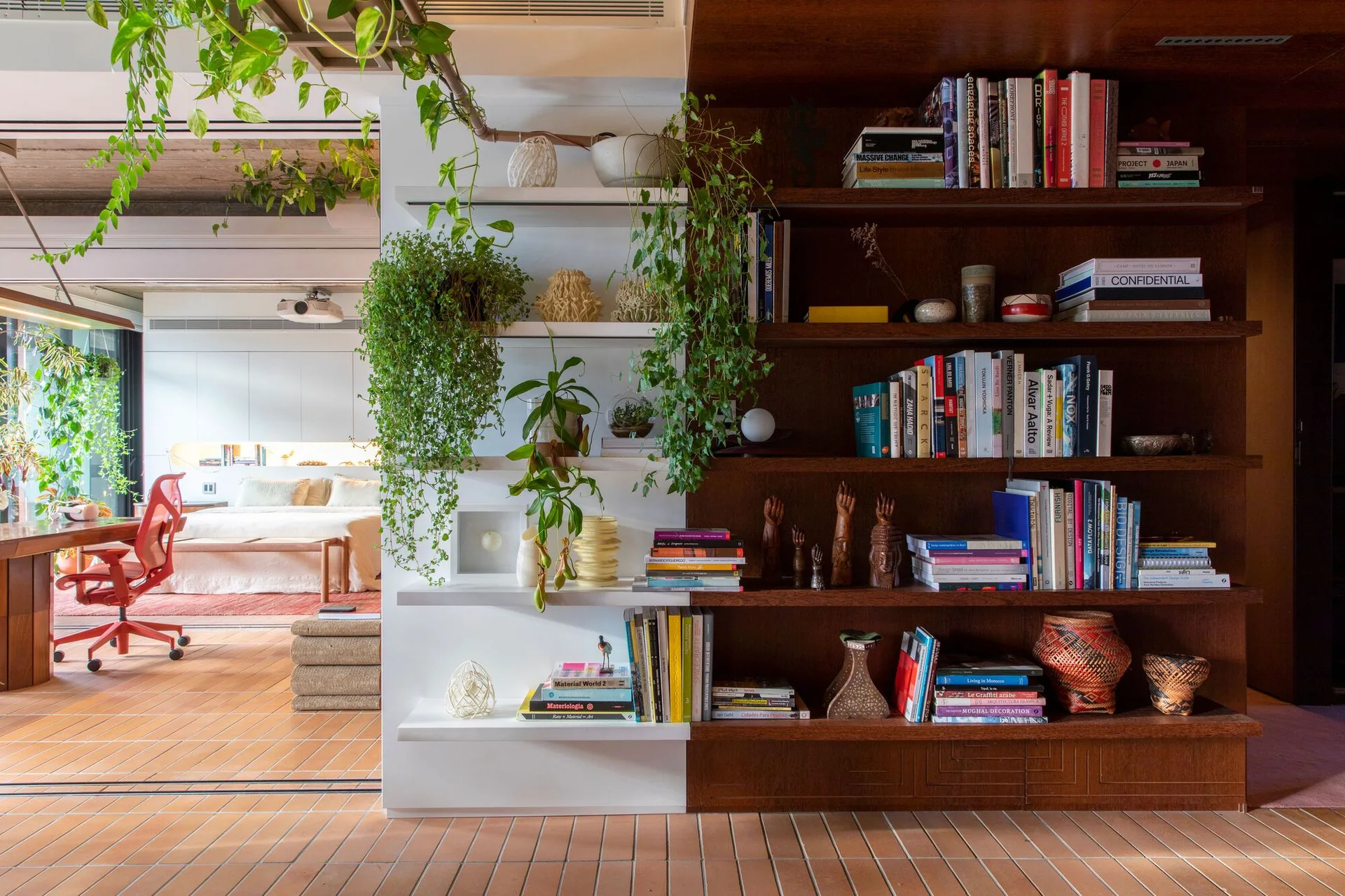 A dual-toned white and dark wood shelving unit filled with books and trailing plants divides a room with terracotta tile flooring.