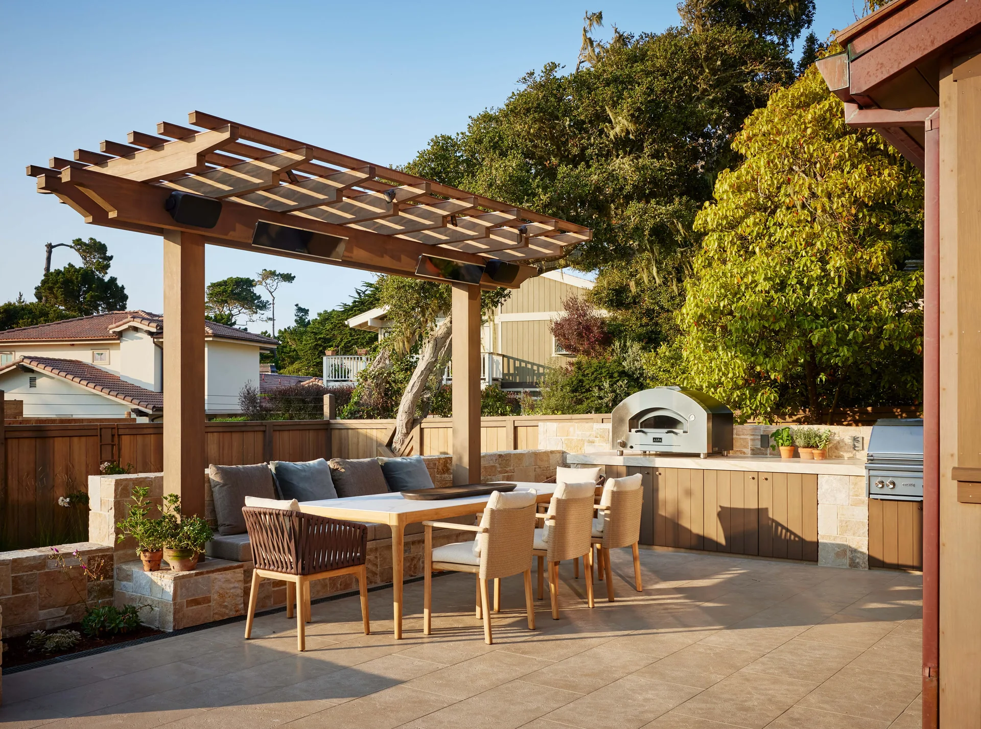 A stone-clad outdoor patio features a wooden dining table and chairs under a pergola next to an integrated kitchen with a built-in pizza oven.