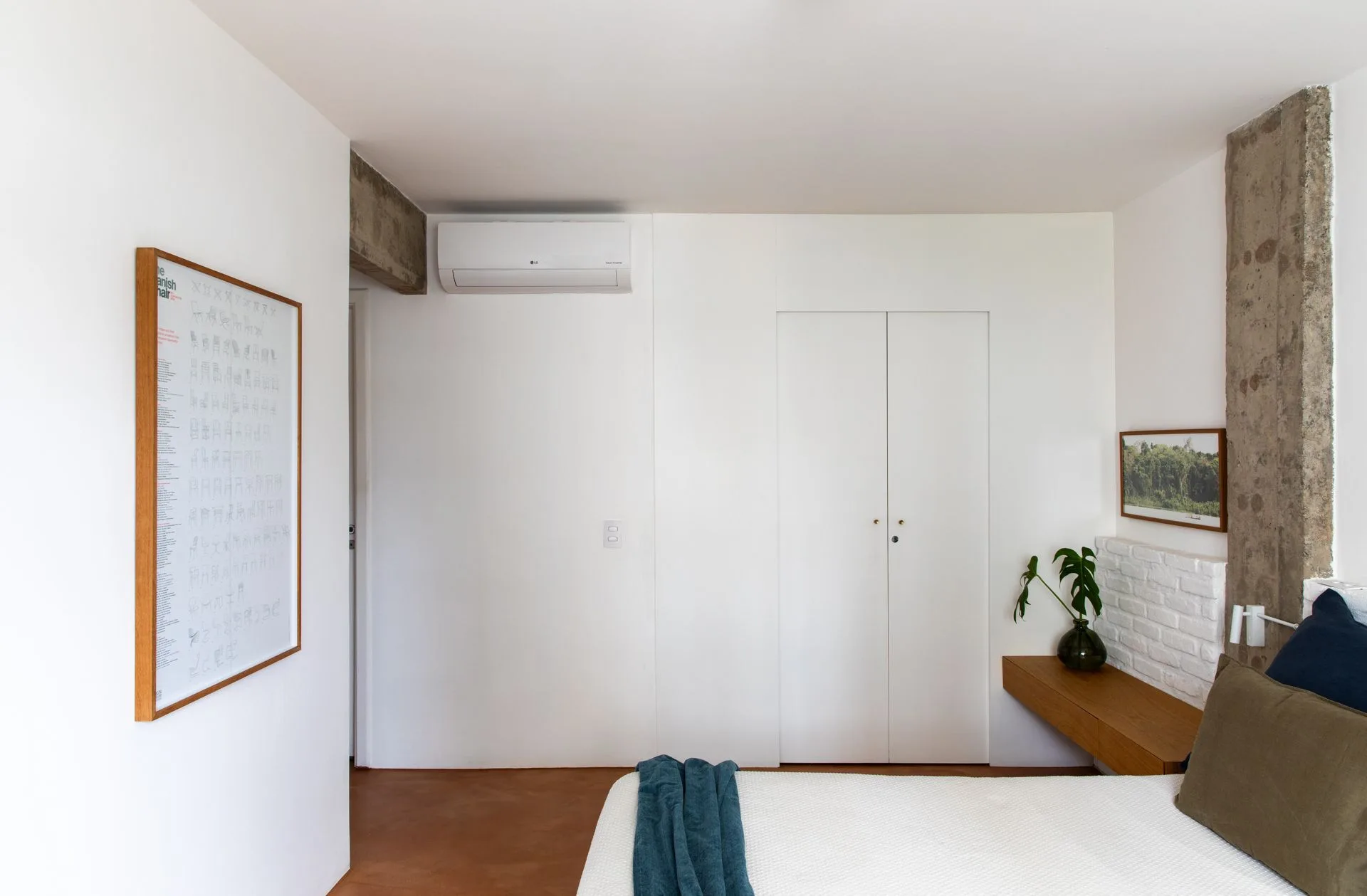 A minimalist bedroom features a bed with blue and olive green pillows, a floating wood shelf, white closet doors, and an exposed concrete column.