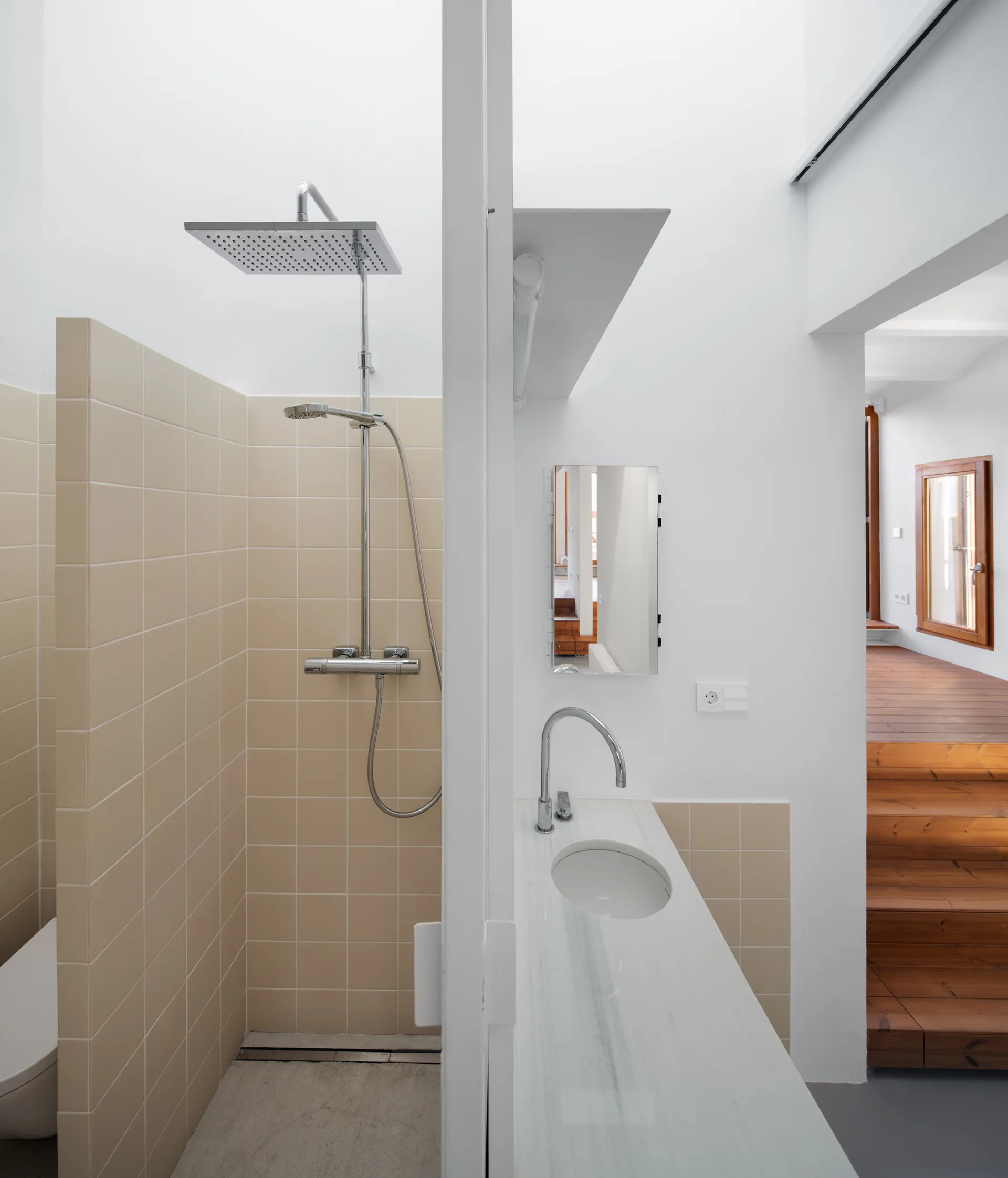 A minimalist bathroom with beige tiled shower walls, a white stone vanity sink, and a view of a wooden staircase in the background.