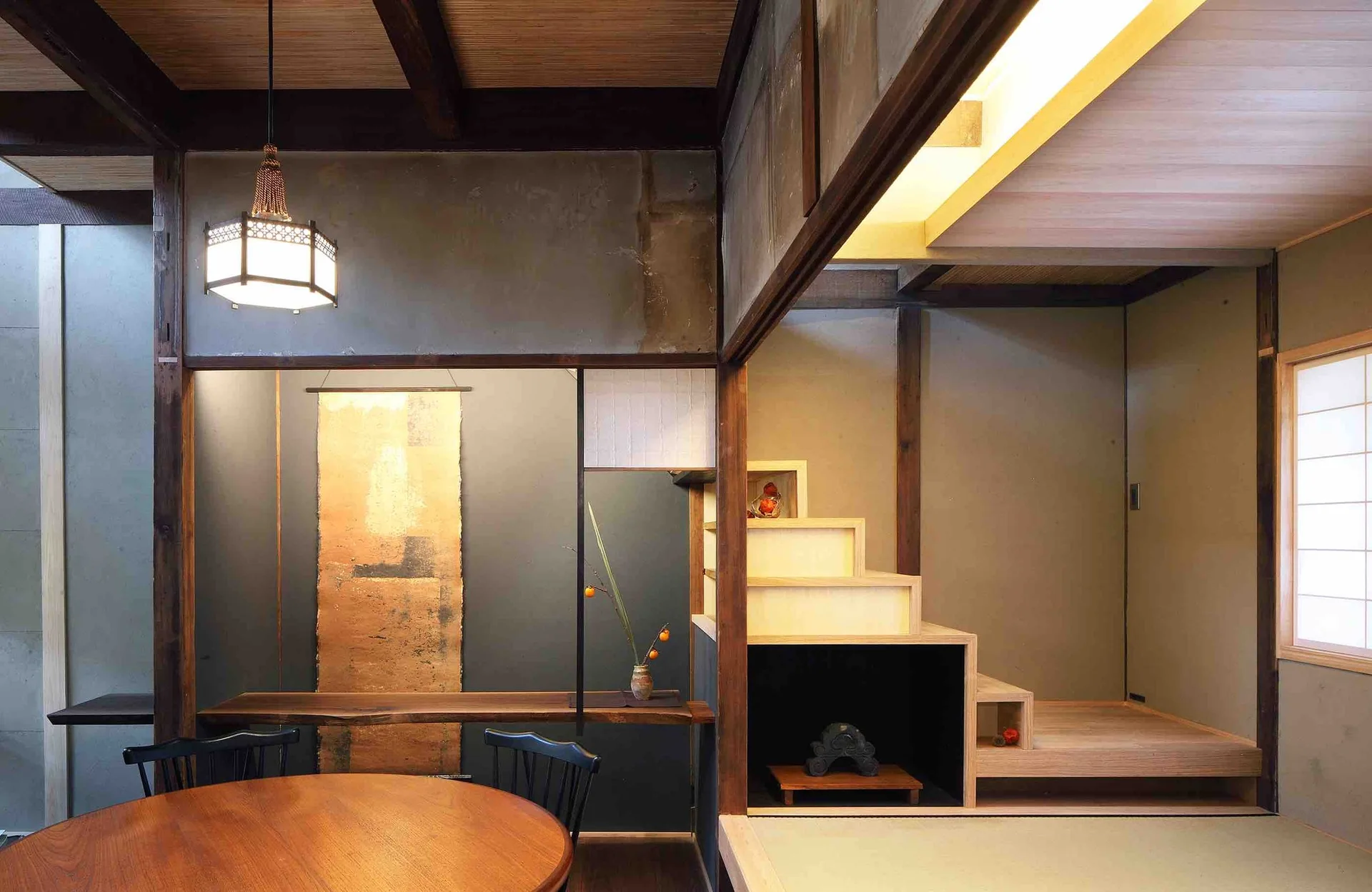 A traditional Japanese dining room with a round wooden table, a pendant lantern, and a wooden staircase featuring integrated storage niches.