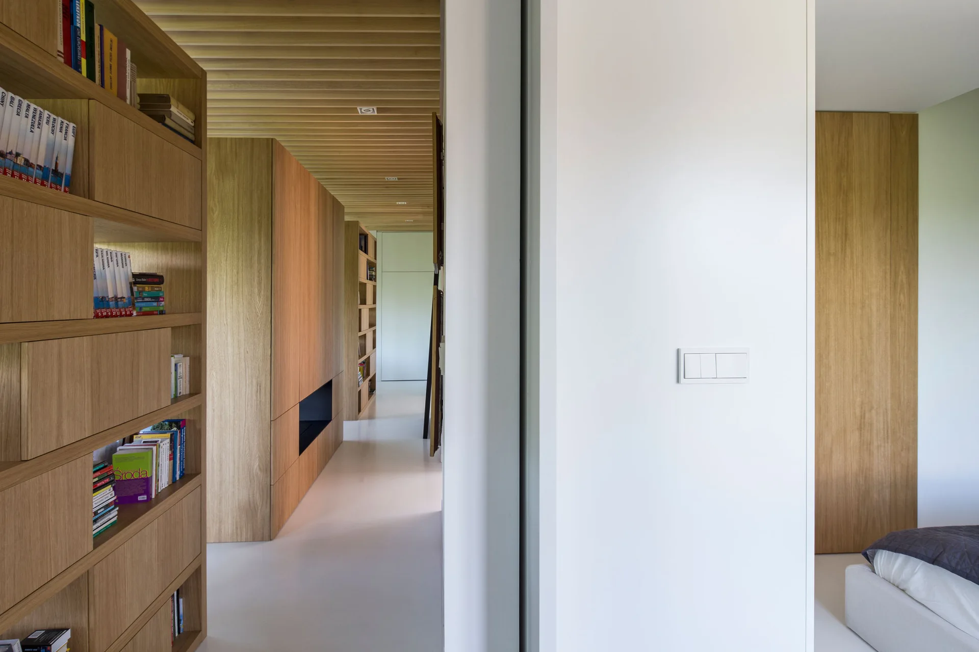 A modern hallway features a wooden slatted ceiling, built-in oak bookshelves, and smooth white flooring adjacent to a minimalist bedroom.