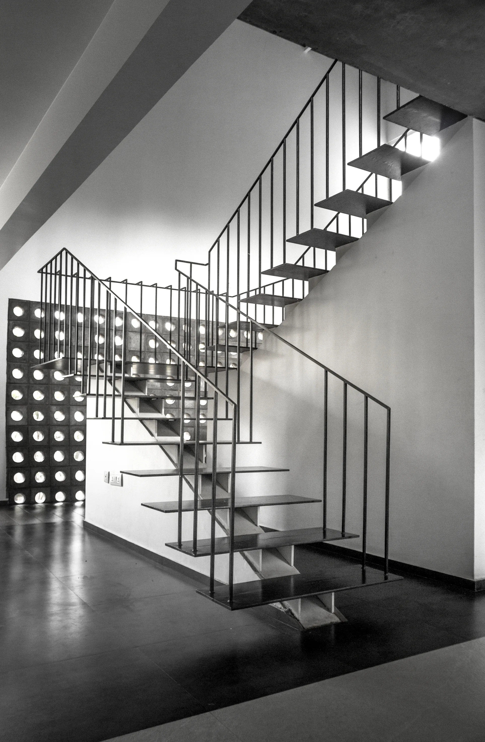 A minimalist floating staircase with black metal treads and railings stands near a white wall and a decorative breeze block screen.