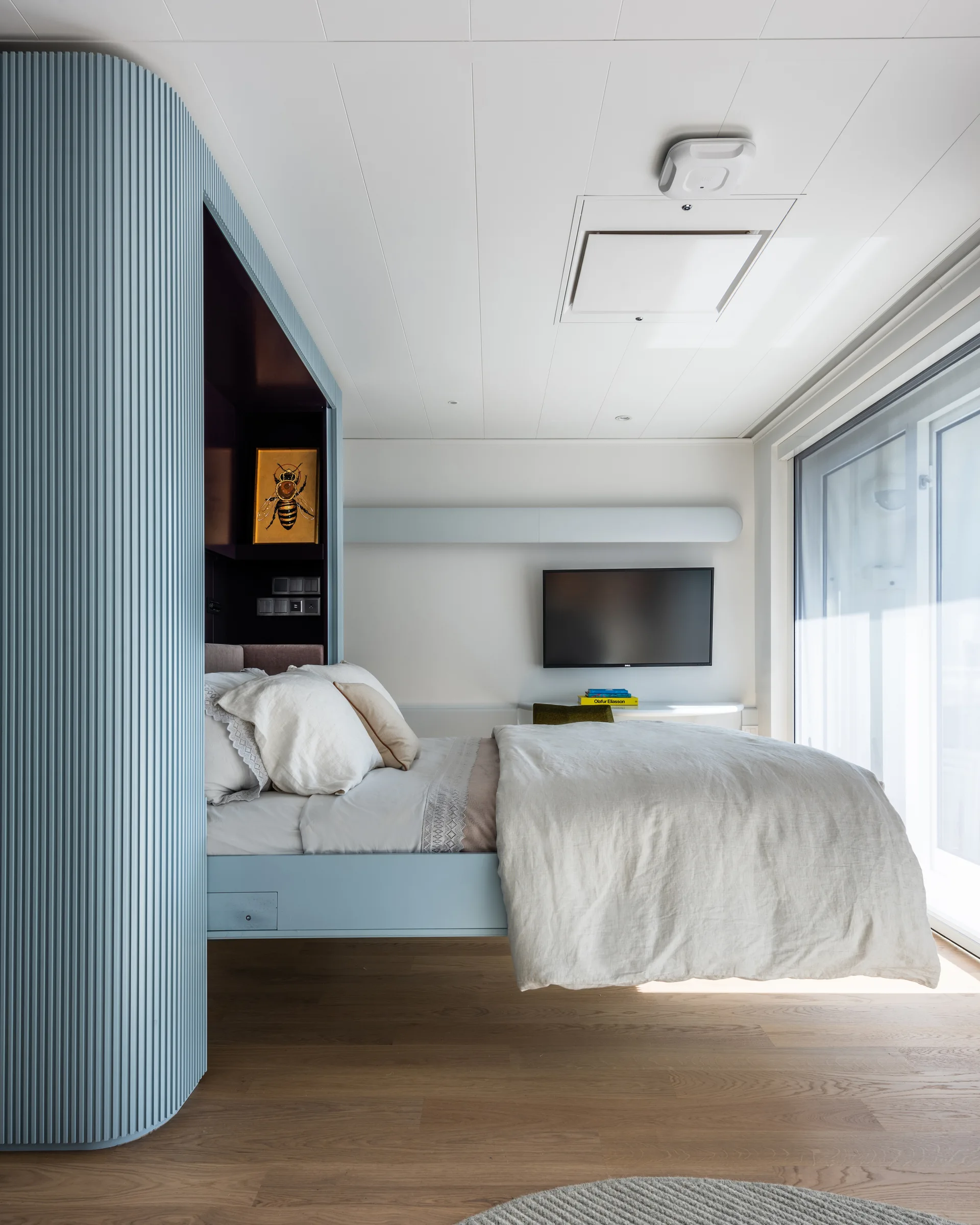 A contemporary bedroom features a floating wood bed frame positioned next to a curved, light-blue fluted partition wall and a large window.