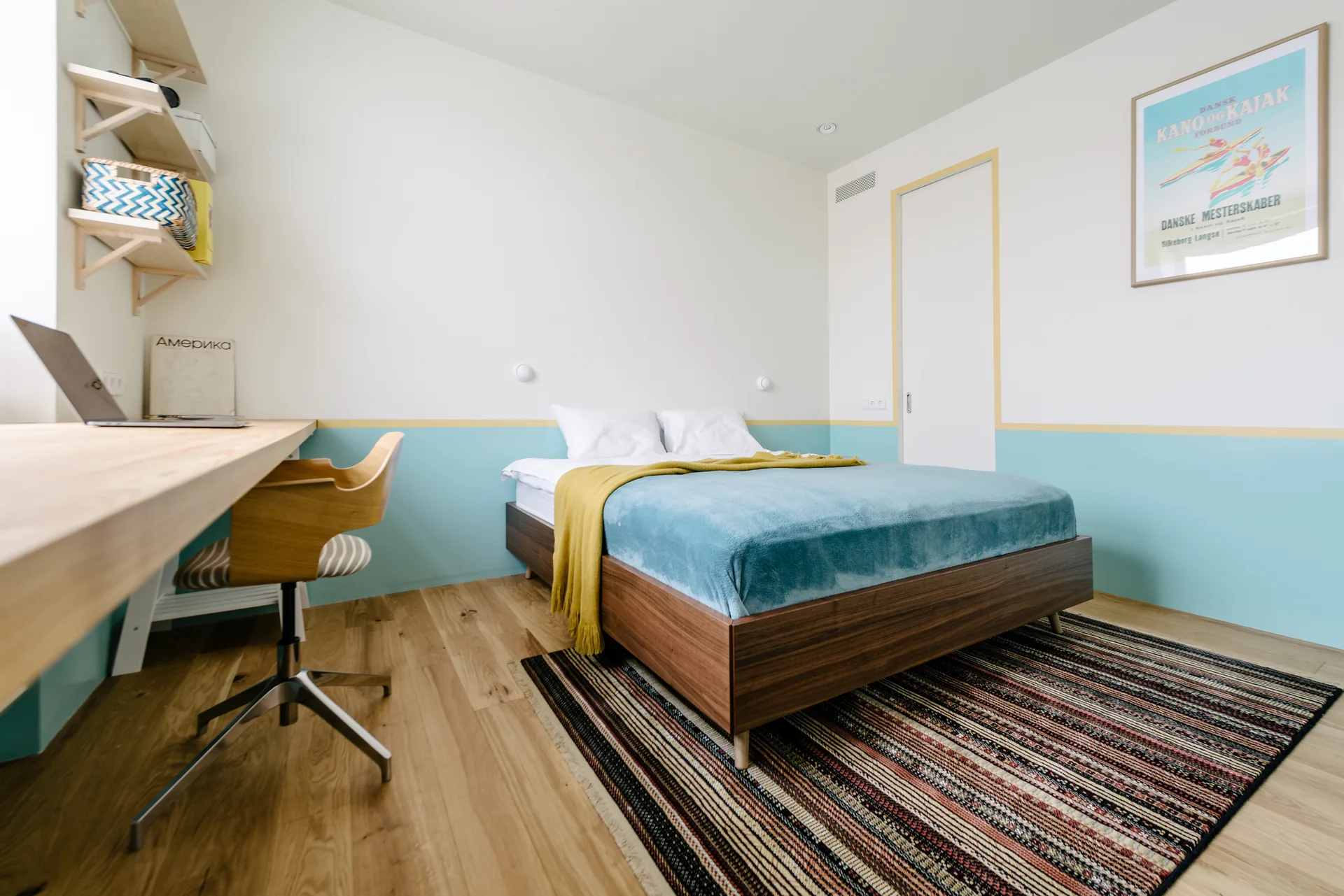 A modern bedroom features a dark wood bed on a striped rug, a light wood built-in desk, and two-tone turquoise and white walls.