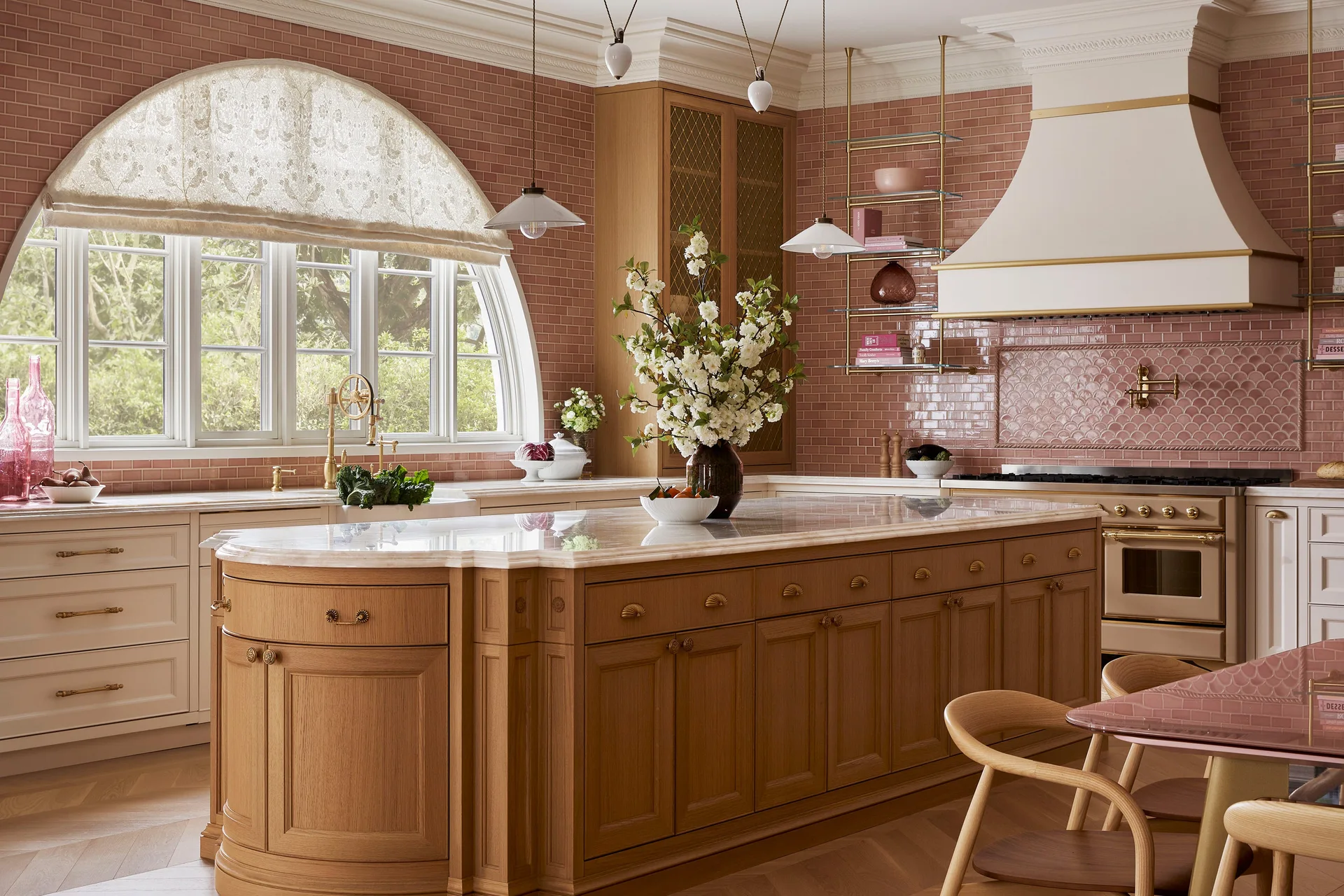 A kitchen features a large curved oak island with a stone countertop, pink subway tile walls, a professional range hood, and a large arched window.