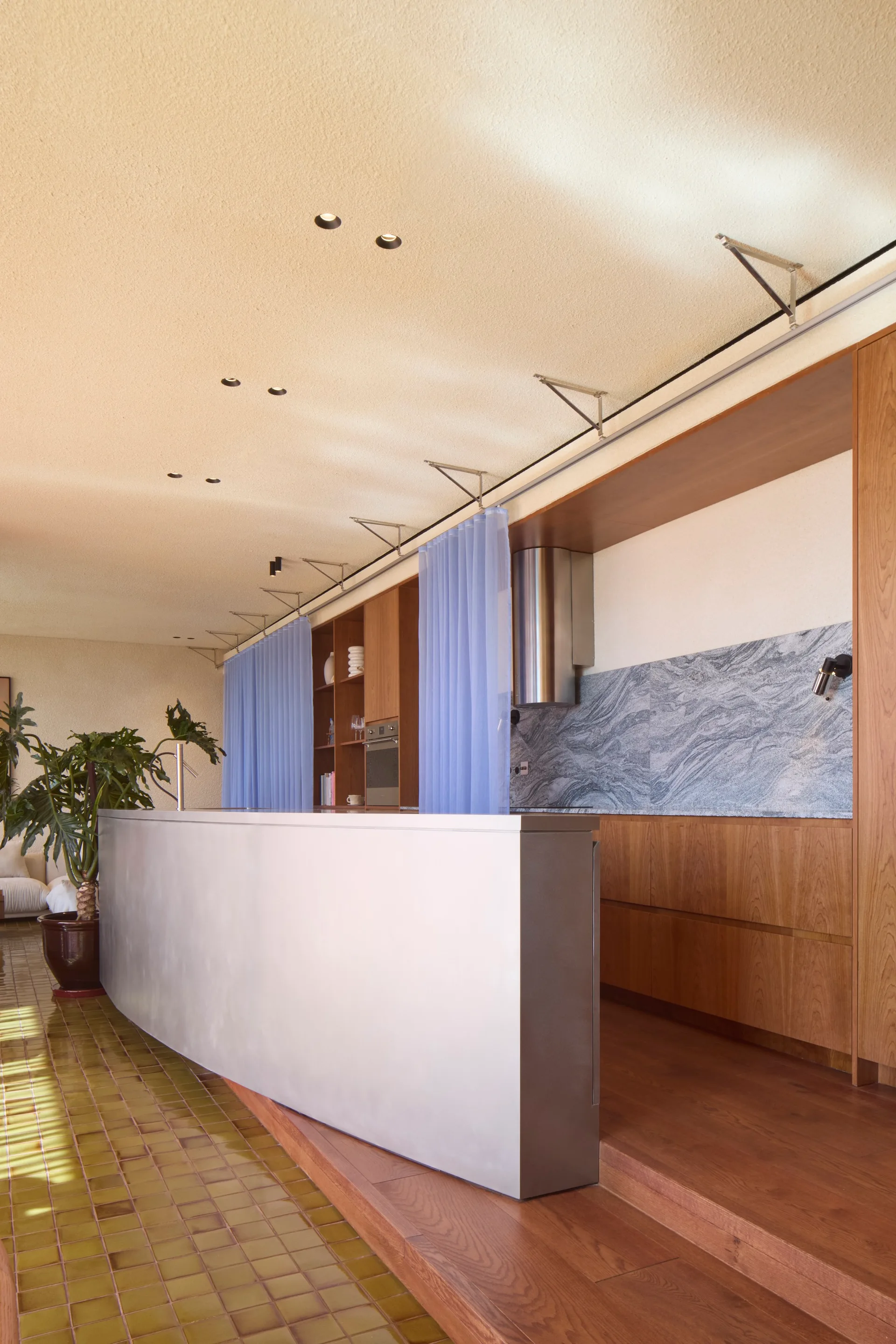 A modern kitchen featuring a brushed metal island, warm wood cabinetry, a grey marble backsplash, and hanging blue sheer curtains.