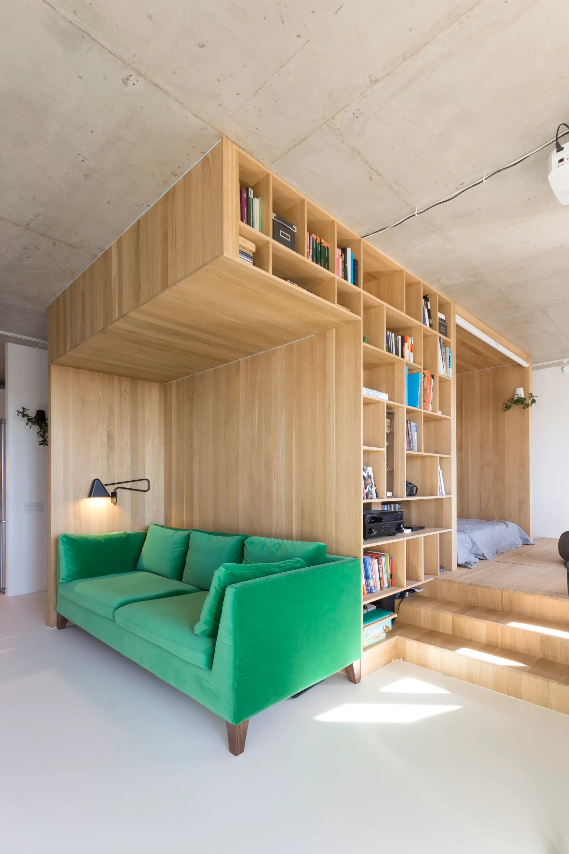 A bright green sofa sits in a niche under a large, custom light wood storage and platform structure against an exposed concrete ceiling.