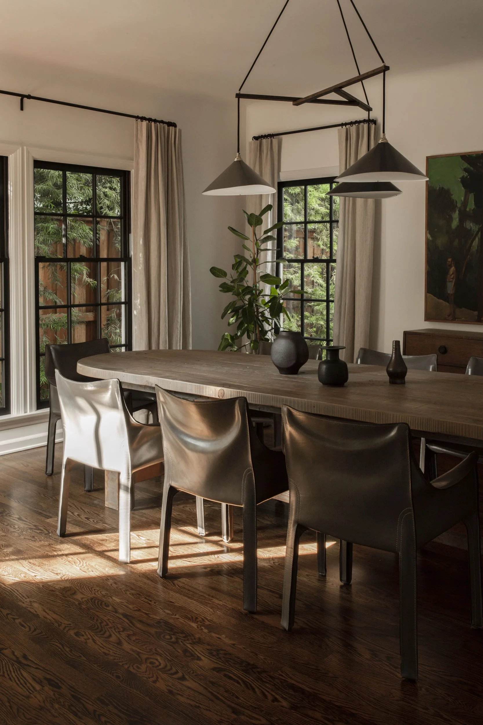 A rustic wooden dining table is surrounded by dark leather chairs under a black multi-cone pendant light in a room with black grid windows.