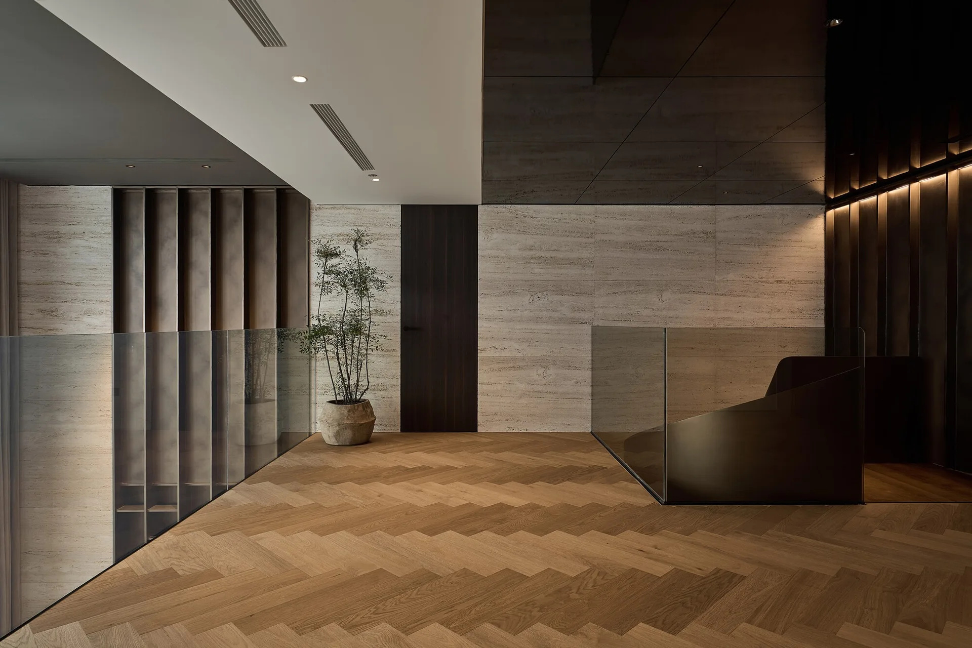 A modern landing featuring herringbone wood flooring, a glass railing, beige stone-clad walls, a dark wood door, and a single potted plant.