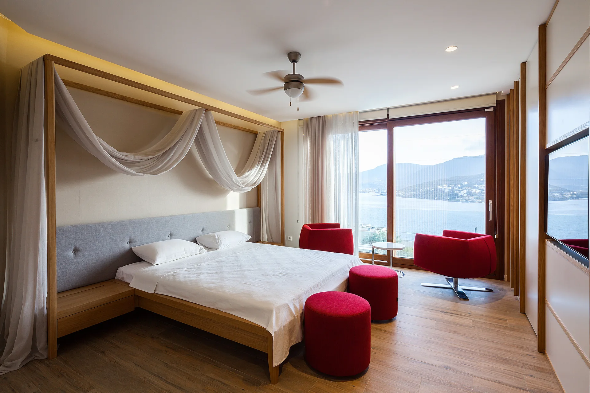 A light wood canopy bed with draped sheer white fabric sits in a bedroom next to bright red lounge chairs and a large window overlooking the water.
