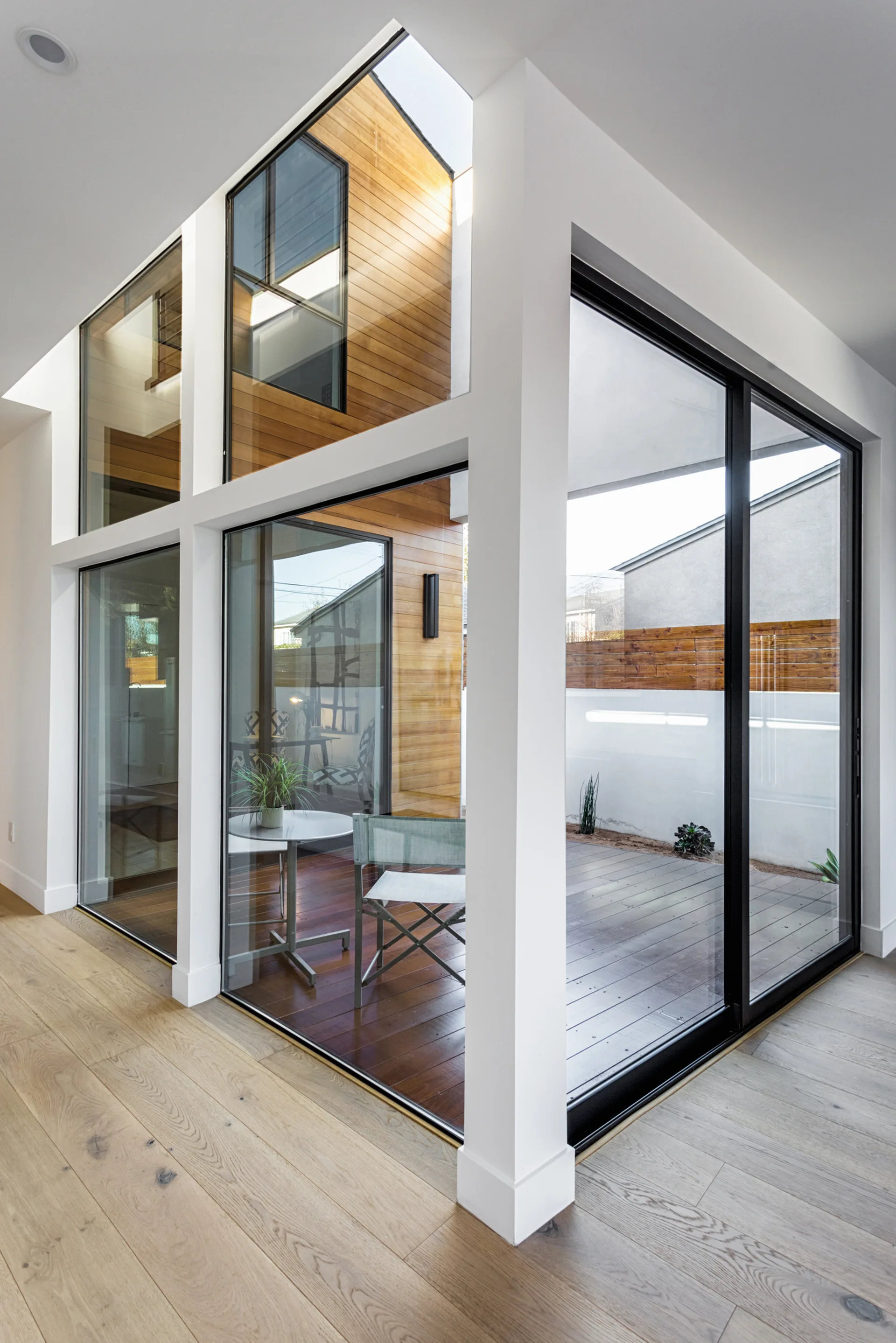 A modern interior with light wood flooring and a large black-framed glass wall looking out onto a wooden deck with a small table and chair.