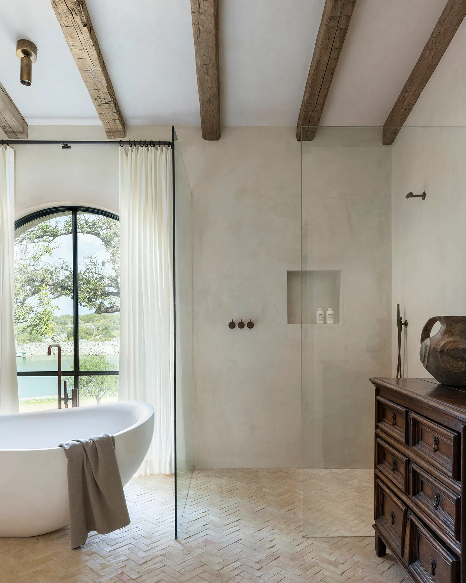 A rustic bathroom with a white freestanding bathtub, frameless glass shower, weathered wood ceiling beams, and herringbone brick flooring.
