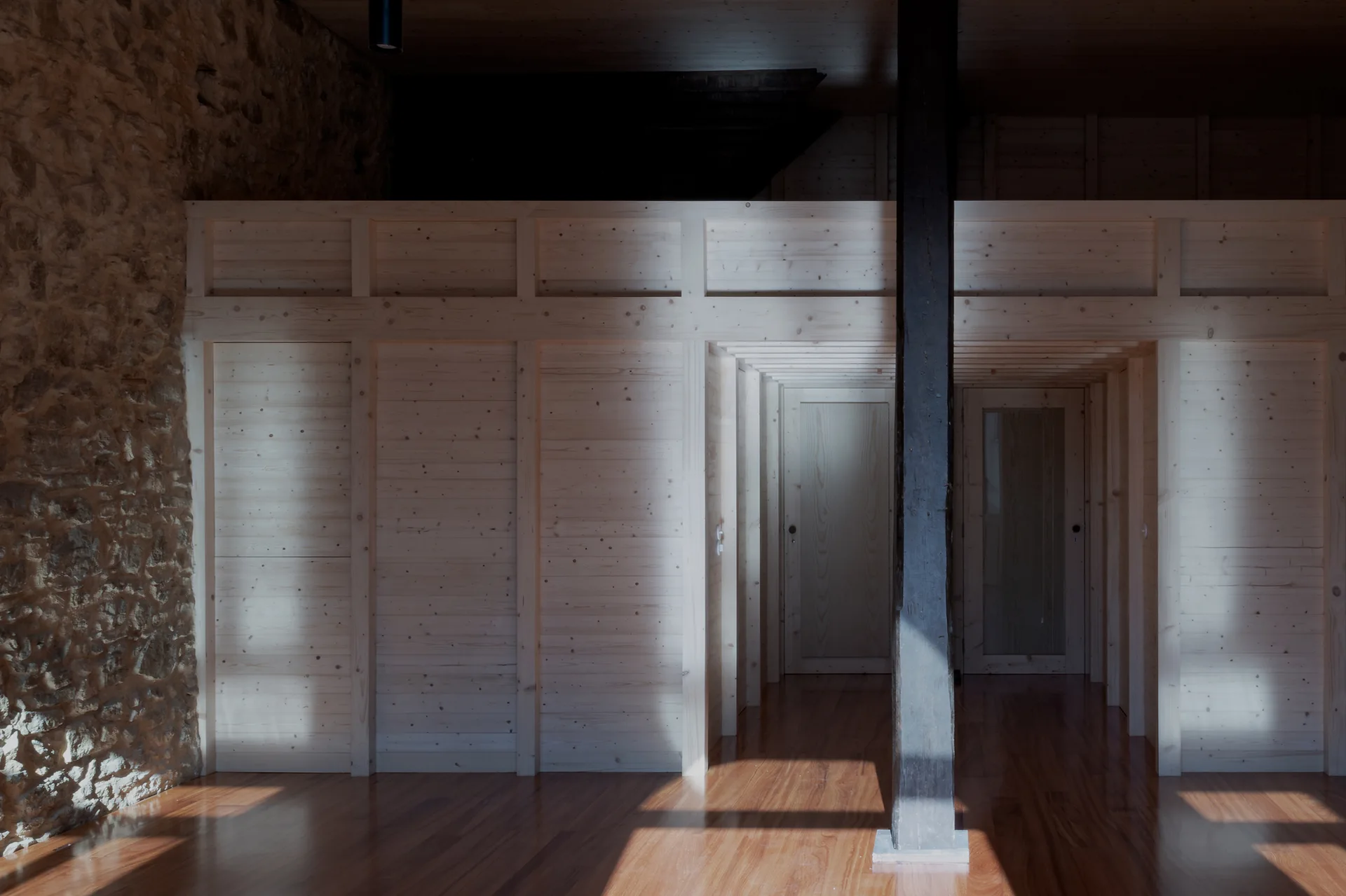 A light pine wood partition wall with doors stands adjacent to a rustic stone wall on a polished hardwood floor.