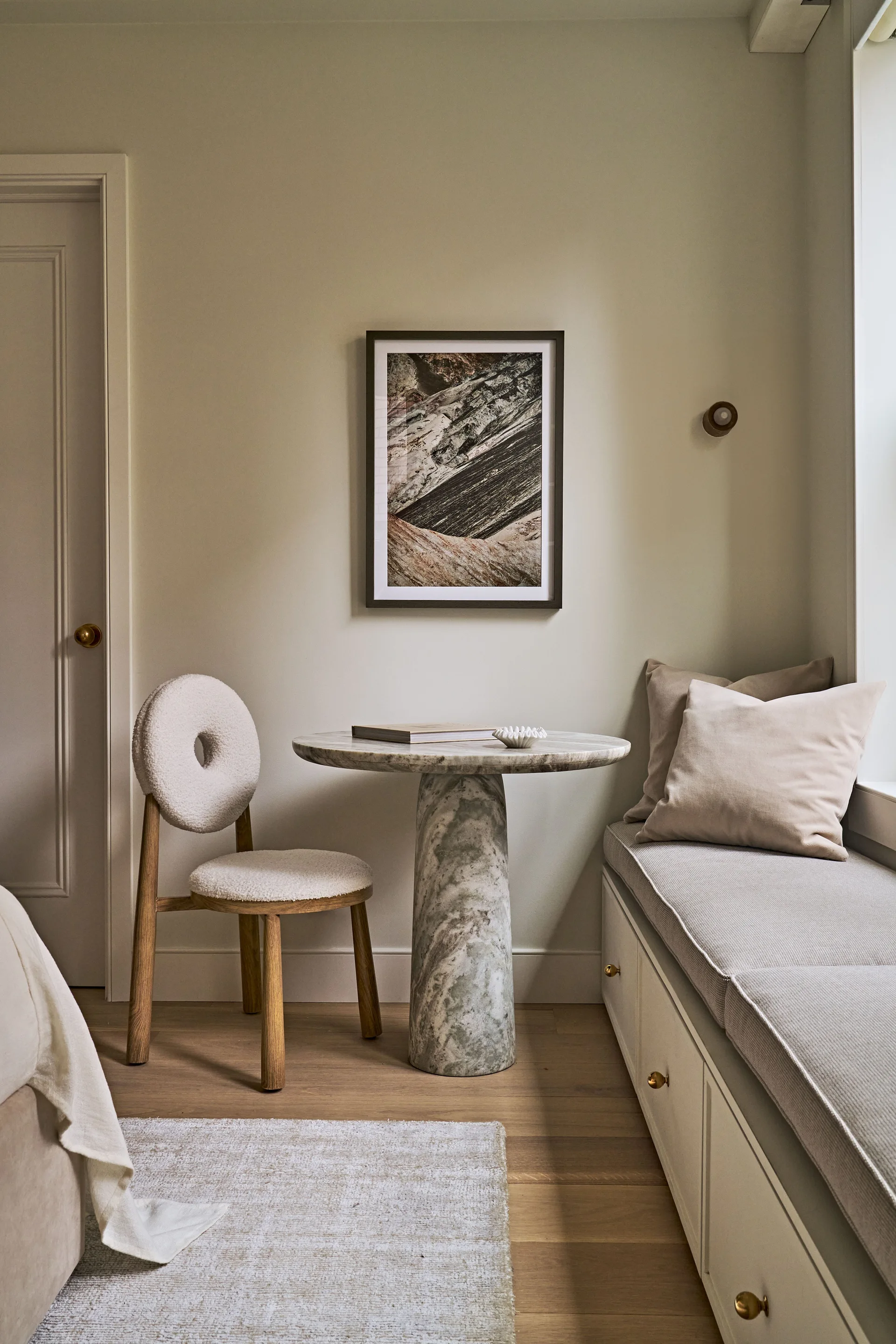 A neutral-toned bedroom corner features a marble pedestal table, a boucle-upholstered chair, and a built-in window bench with storage drawers.