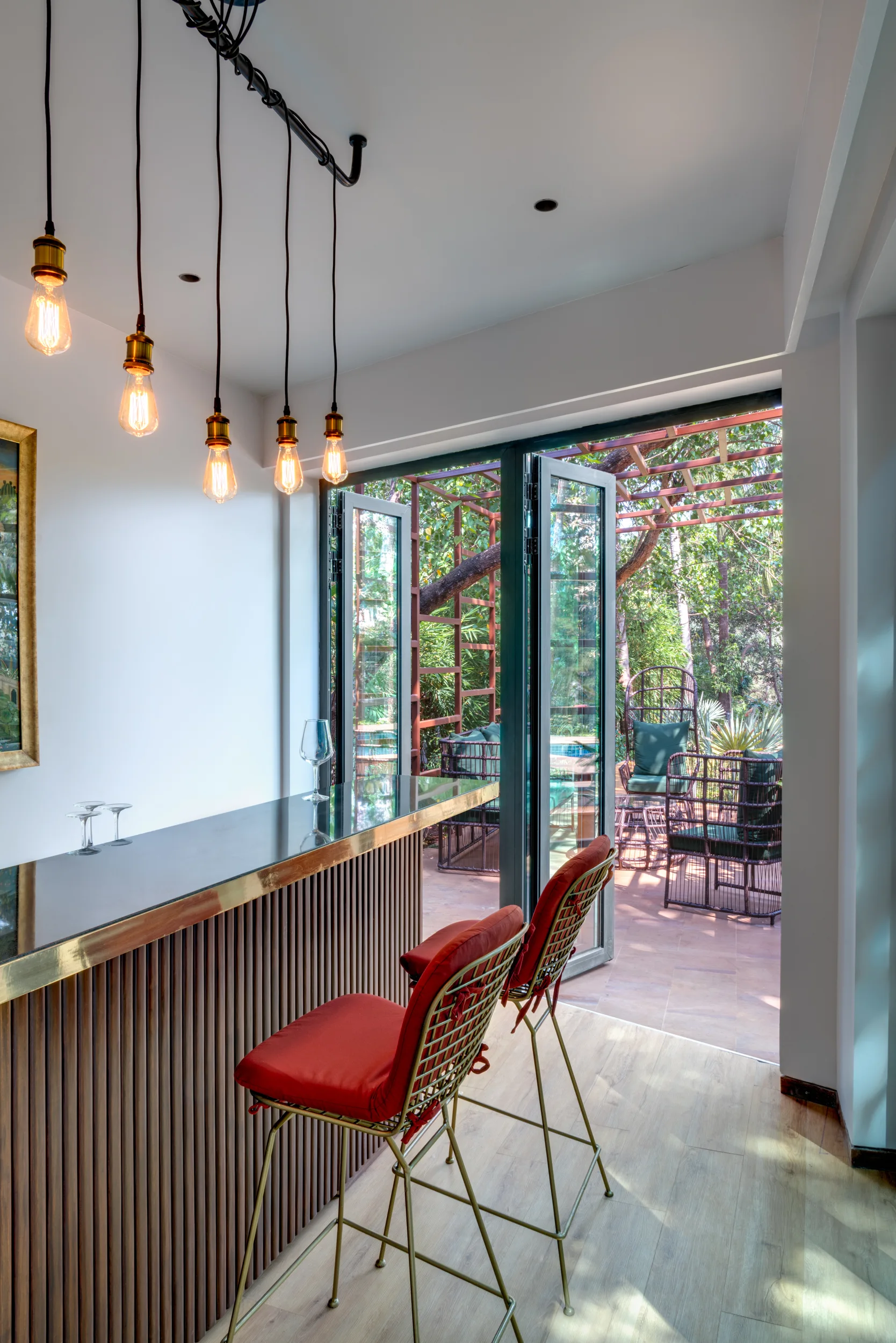 A modern indoor bar with a fluted wood base and red stools stands beside open glass bifold doors opening to a green patio.