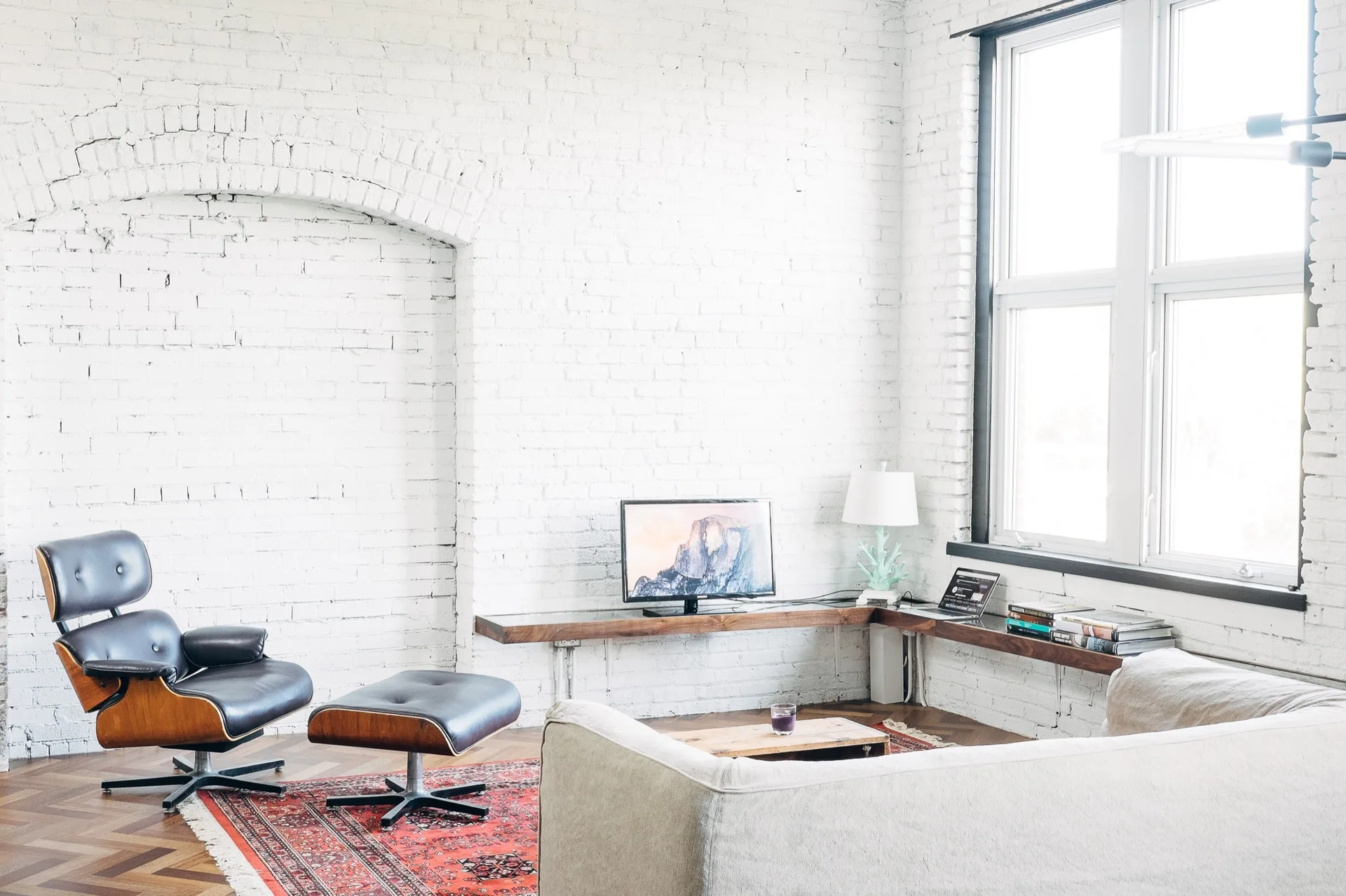 A living room features white painted brick walls, a dark leather lounge chair with ottoman, a red patterned rug, and a floating wood corner shelf.