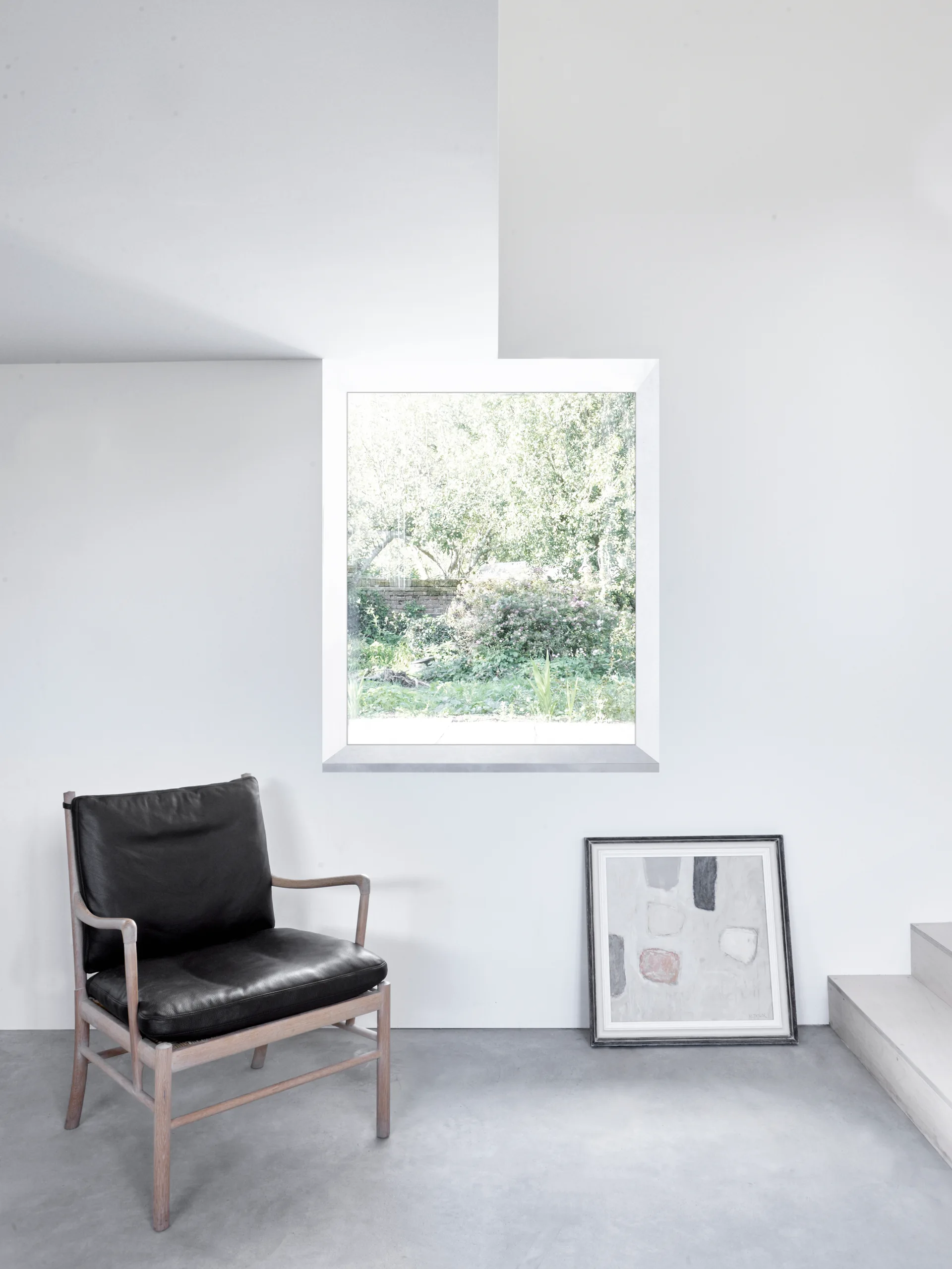 A minimalist room features a black leather armchair, a framed painting resting on a gray concrete floor, and a window displaying a garden view.