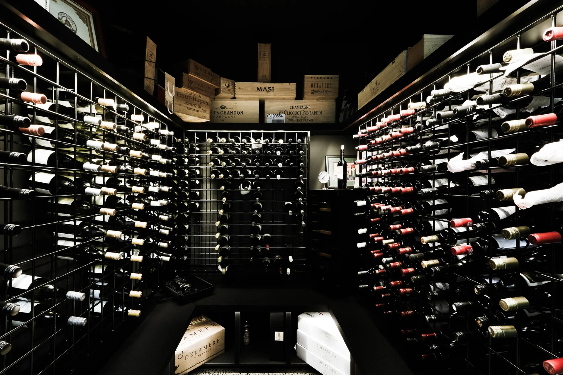 A dark-painted wine cellar features tall black metal grid racks filled with bottles, with wooden wine crates stacked on the upper shelves.