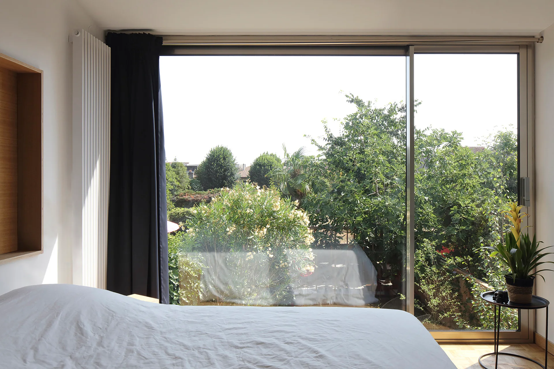 A minimalist bedroom with white bedding features a large glass sliding door looking out onto a lush green garden.