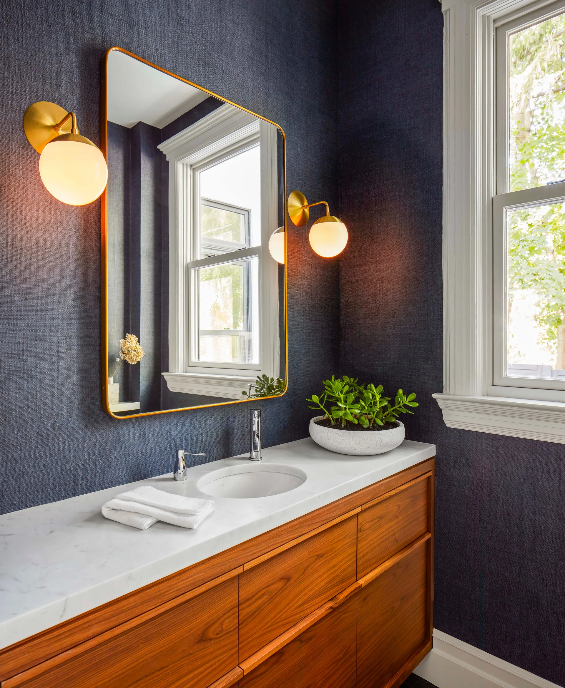A wooden vanity with a white marble countertop is set against navy grasscloth wallpaper under a brass-framed mirror flanked by two globe sconces.