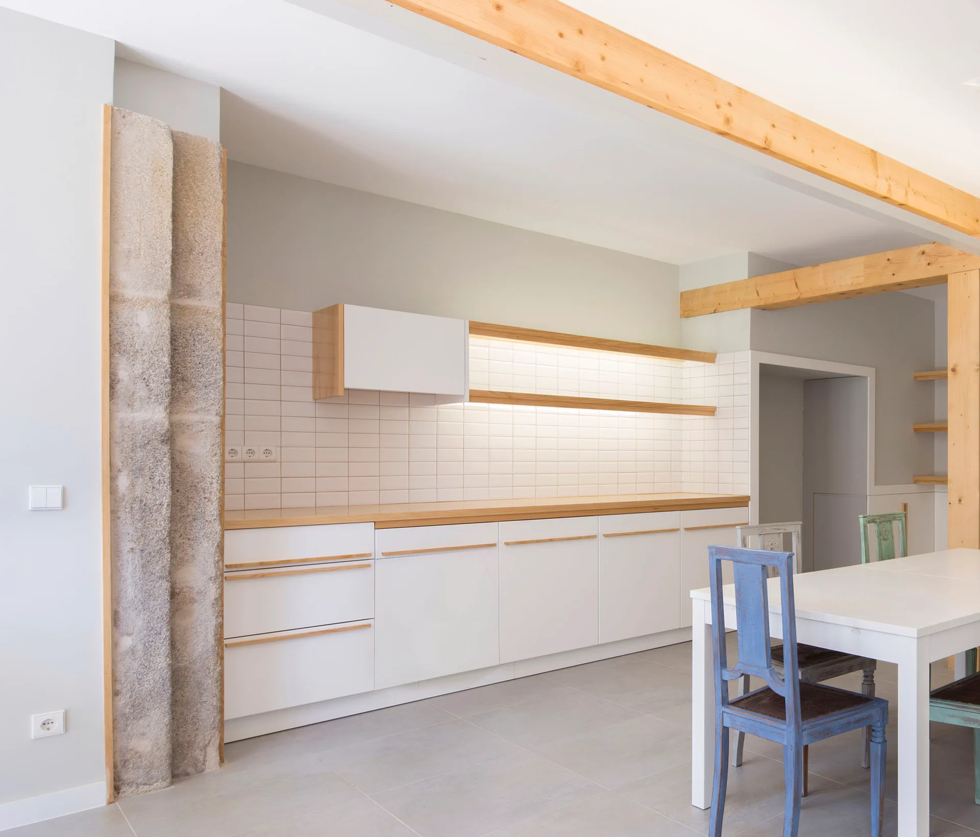 A modern kitchen with white cabinetry, light wood countertops, an exposed ceiling beam, a rough stone pillar, and a dining table.