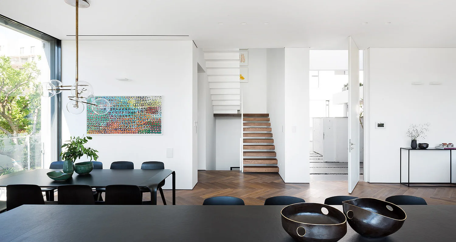 A modern open-plan dining room with black furniture, a colorful art piece, a floating staircase, and dark herringbone wood floors.