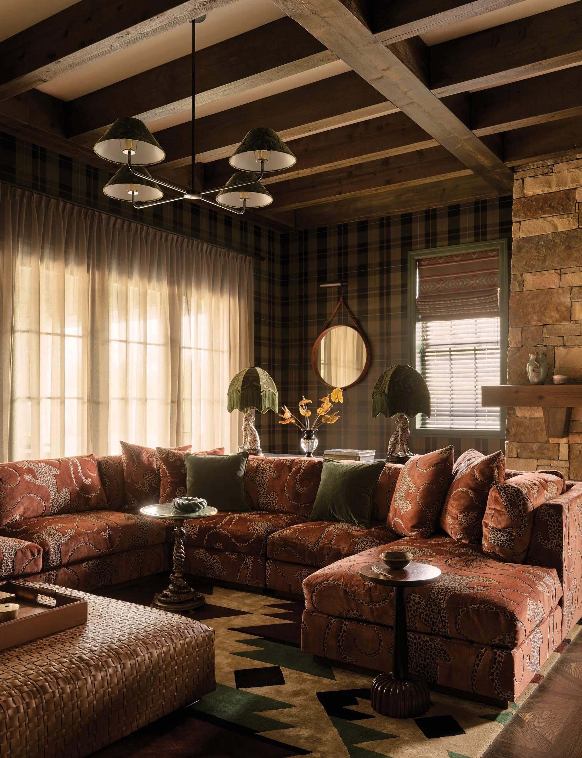A cozy rustic living room featuring a patterned rust sectional, plaid wallpaper, dark wood ceiling beams, and a natural stone fireplace.