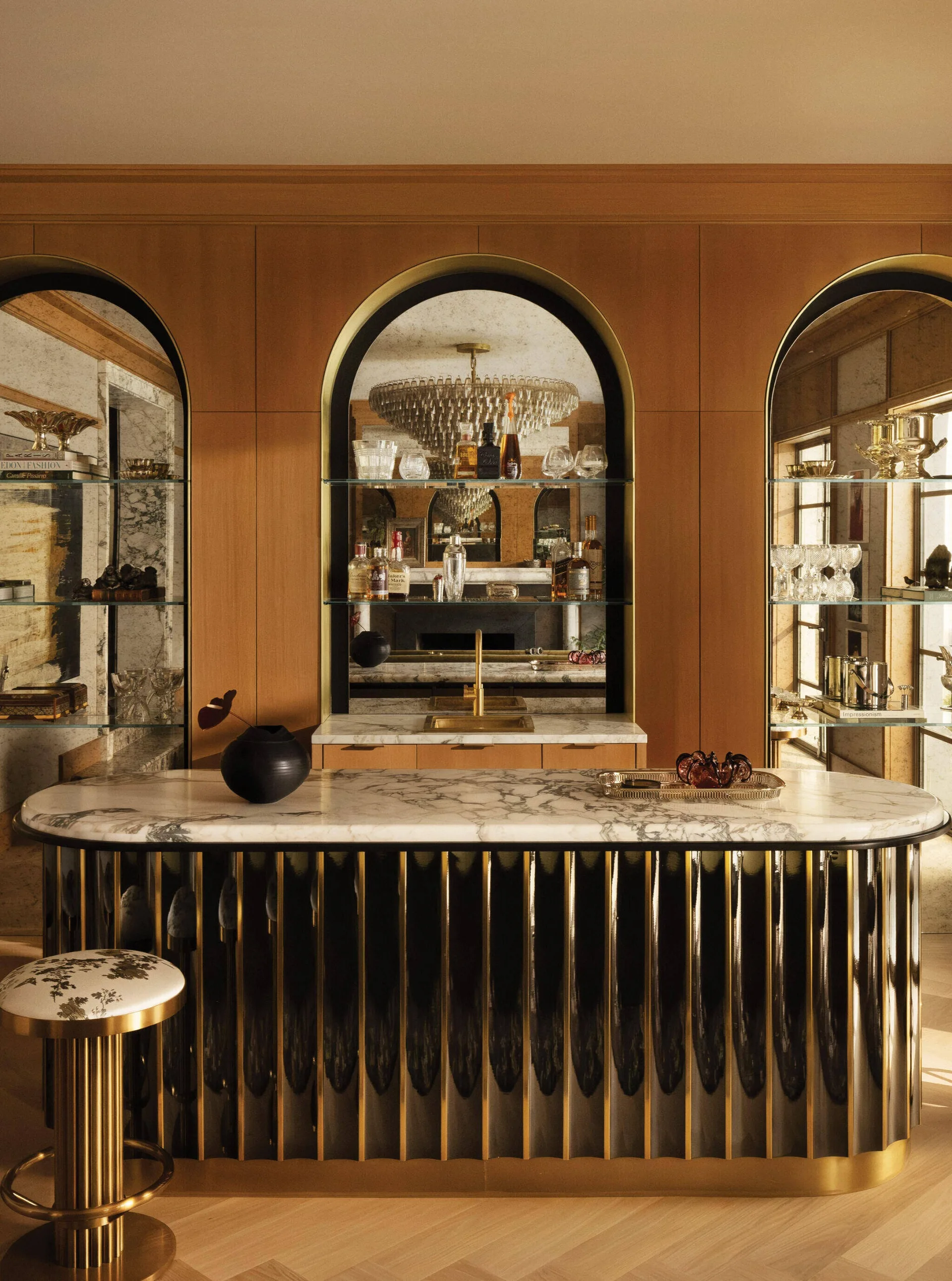 A home bar features a marble-topped island with a fluted black and gold base in front of wood-paneled walls with three arched mirrored display niches.