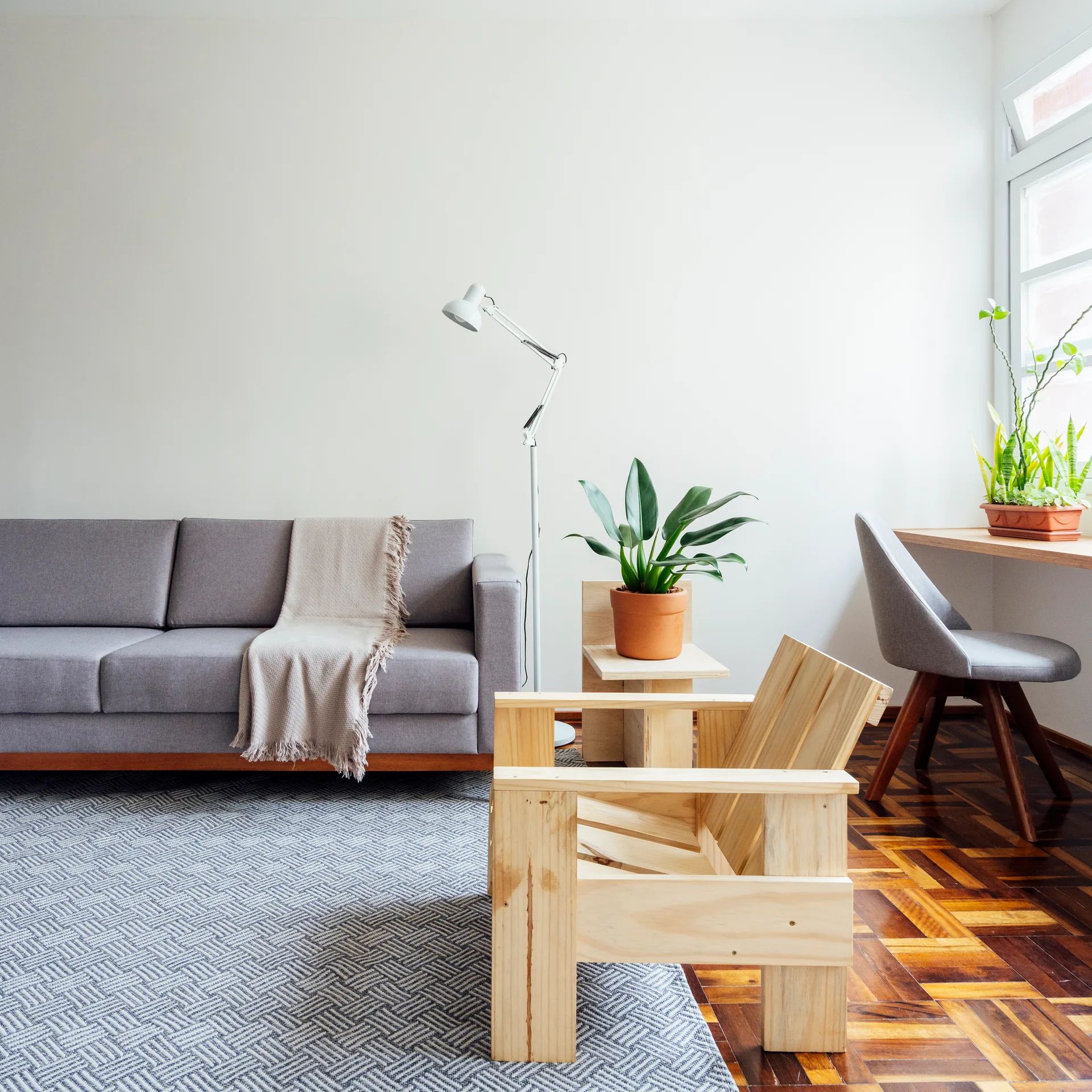 A minimalist living room with a gray sofa, geometric light wood armchair, patterned area rug, and a built-in window desk with a gray chair.
