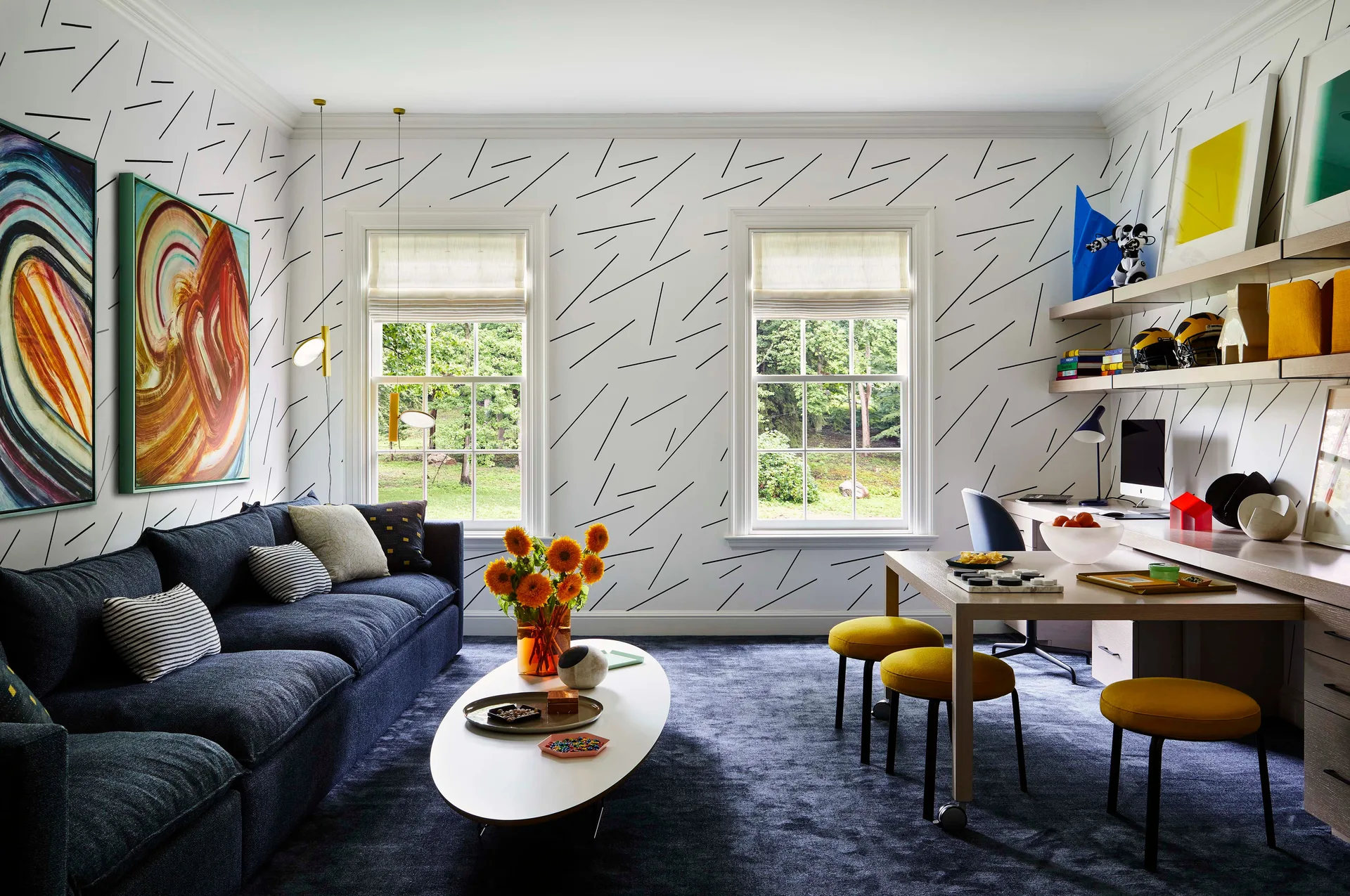A multifunctional room featuring a blue sofa, graphic line-patterned wallpaper, and a home office desk with floating shelves and yellow stools.