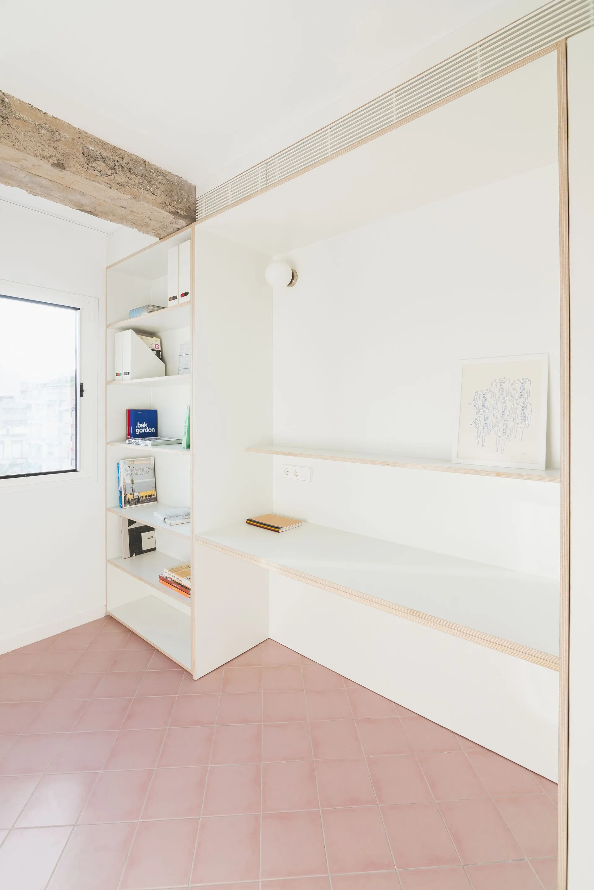 A minimalist workspace featuring a custom white built-in desk and shelves with plywood edges, an exposed ceiling beam, and pink tiled floors.