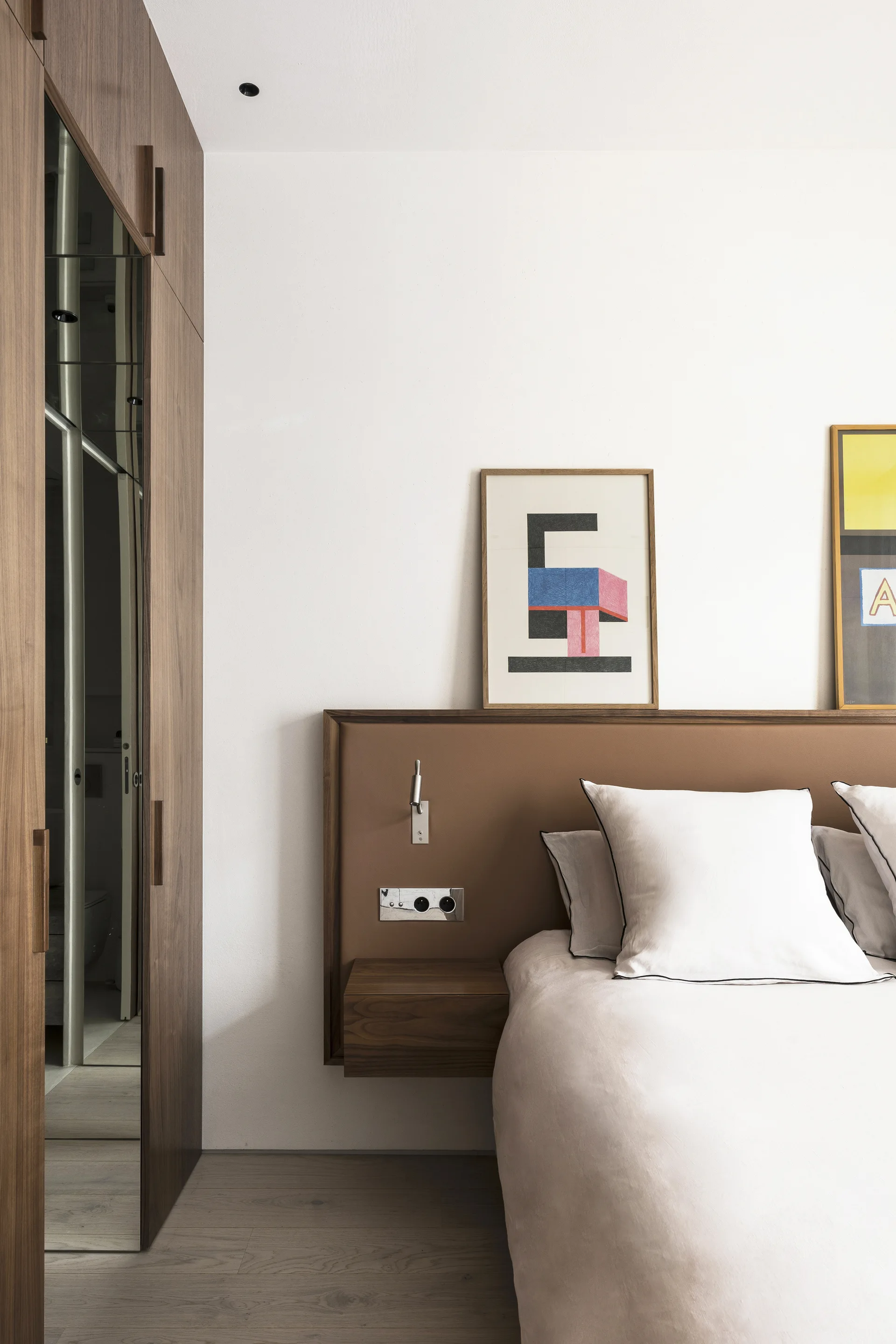 A modern bedroom featuring a wood-paneled headboard with an integrated floating drawer next to a built-in wardrobe with a mirrored door.