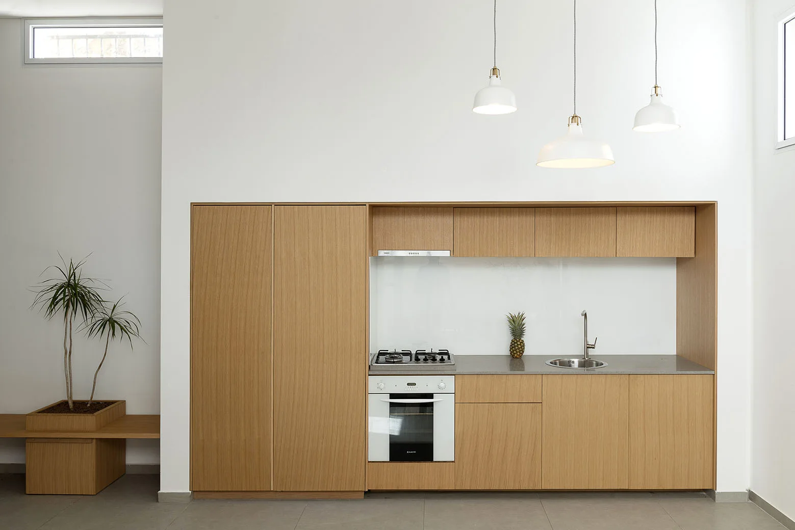A minimalist kitchen features flat-front wood cabinetry recessed into a smooth white wall beneath three hanging white pendant lights.