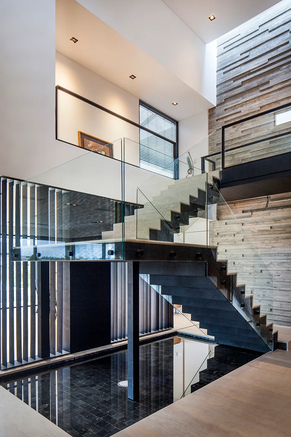 A modern floating staircase with glass railings rises above a black tiled indoor water feature alongside a textured grey stone wall.