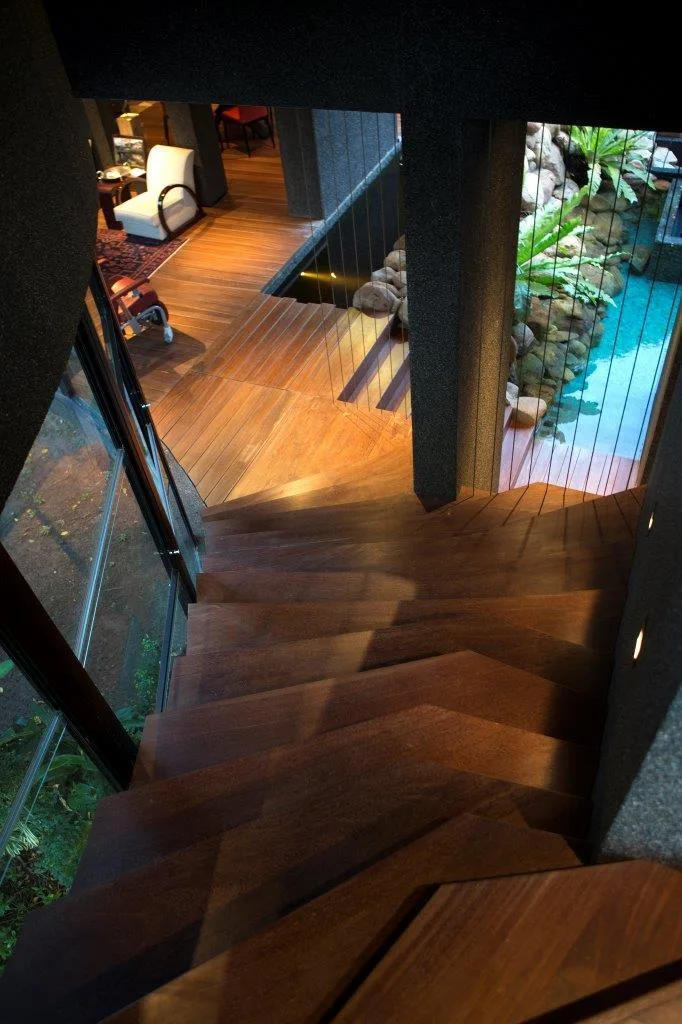 High-angle view of a dark wood staircase descending to a matching wood floor next to an indoor pond with vertical cable railings.