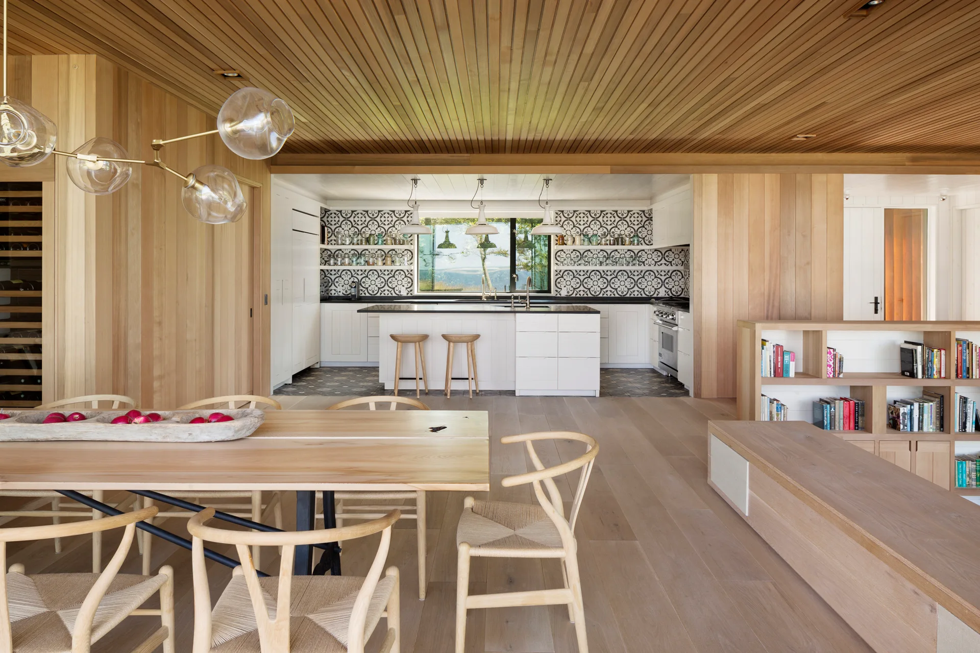 An open-concept dining area with natural wood-paneled walls and ceiling leading to a white kitchen featuring a graphic patterned tile backsplash.
