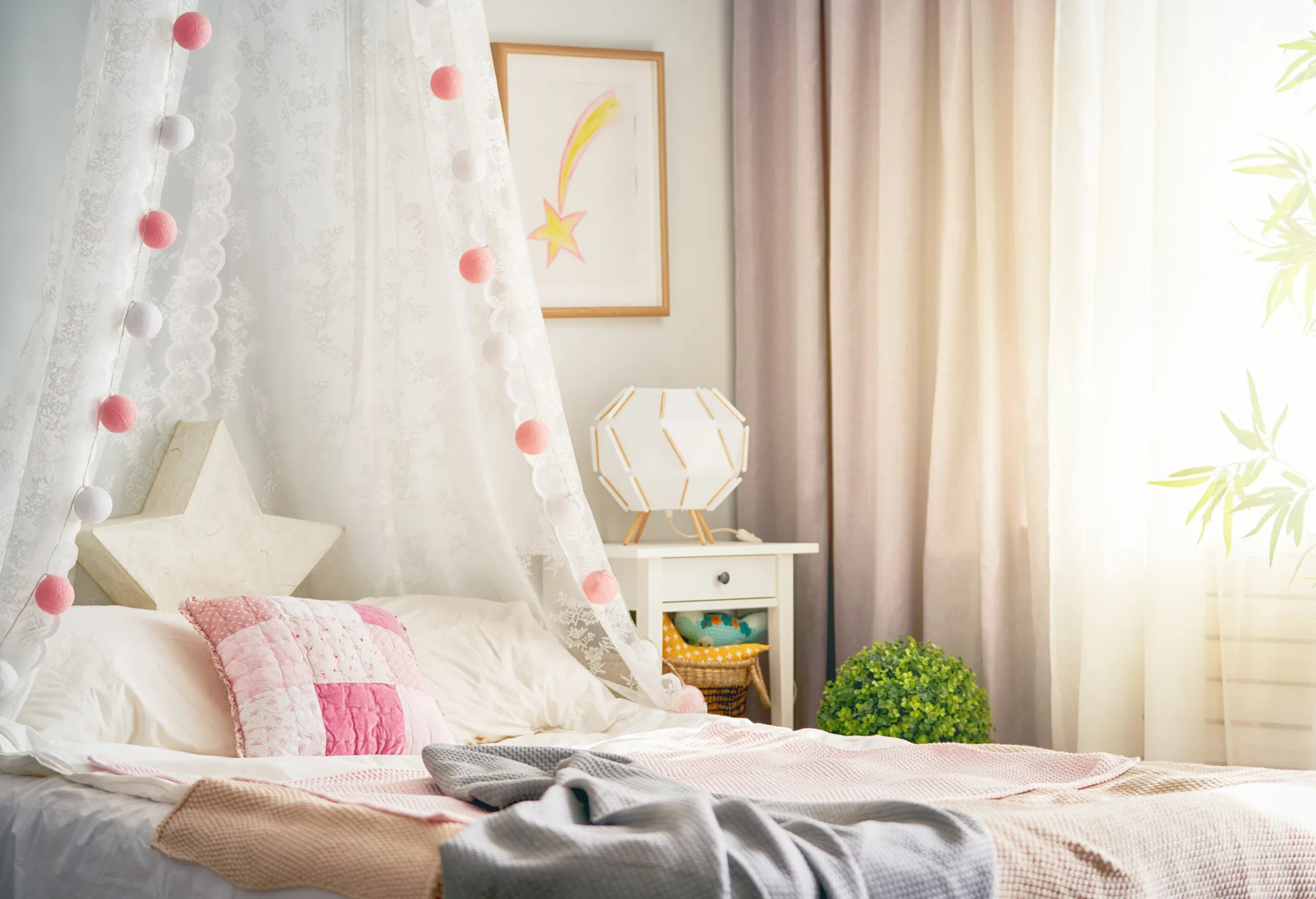 A children's bed features a sheer lace canopy adorned with pink and white ball garland next to a white nightstand with a geometric table lamp.