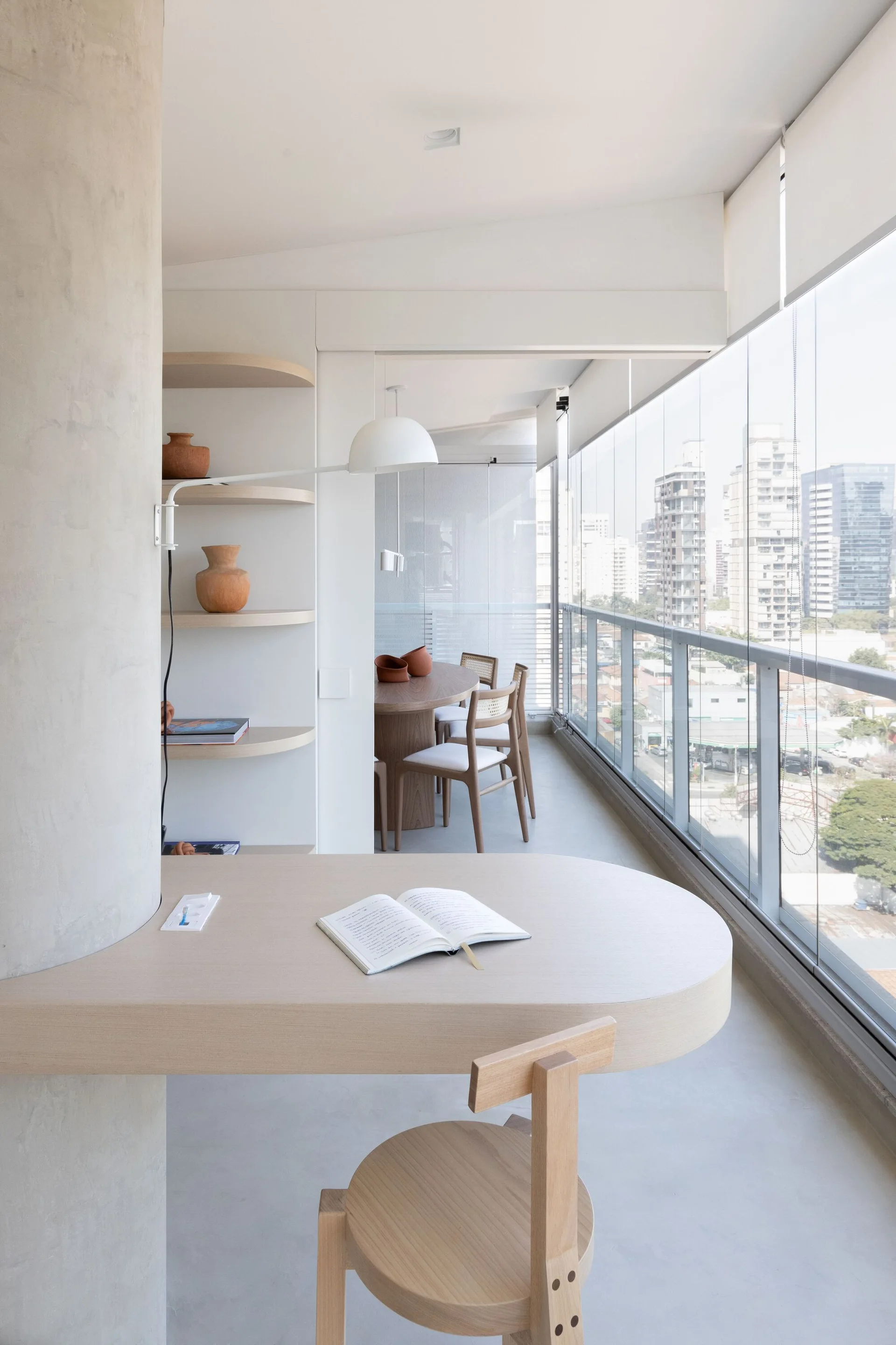 A light wood curved desk is built around a concrete column in a modern apartment beside floor-to-ceiling glass windows.