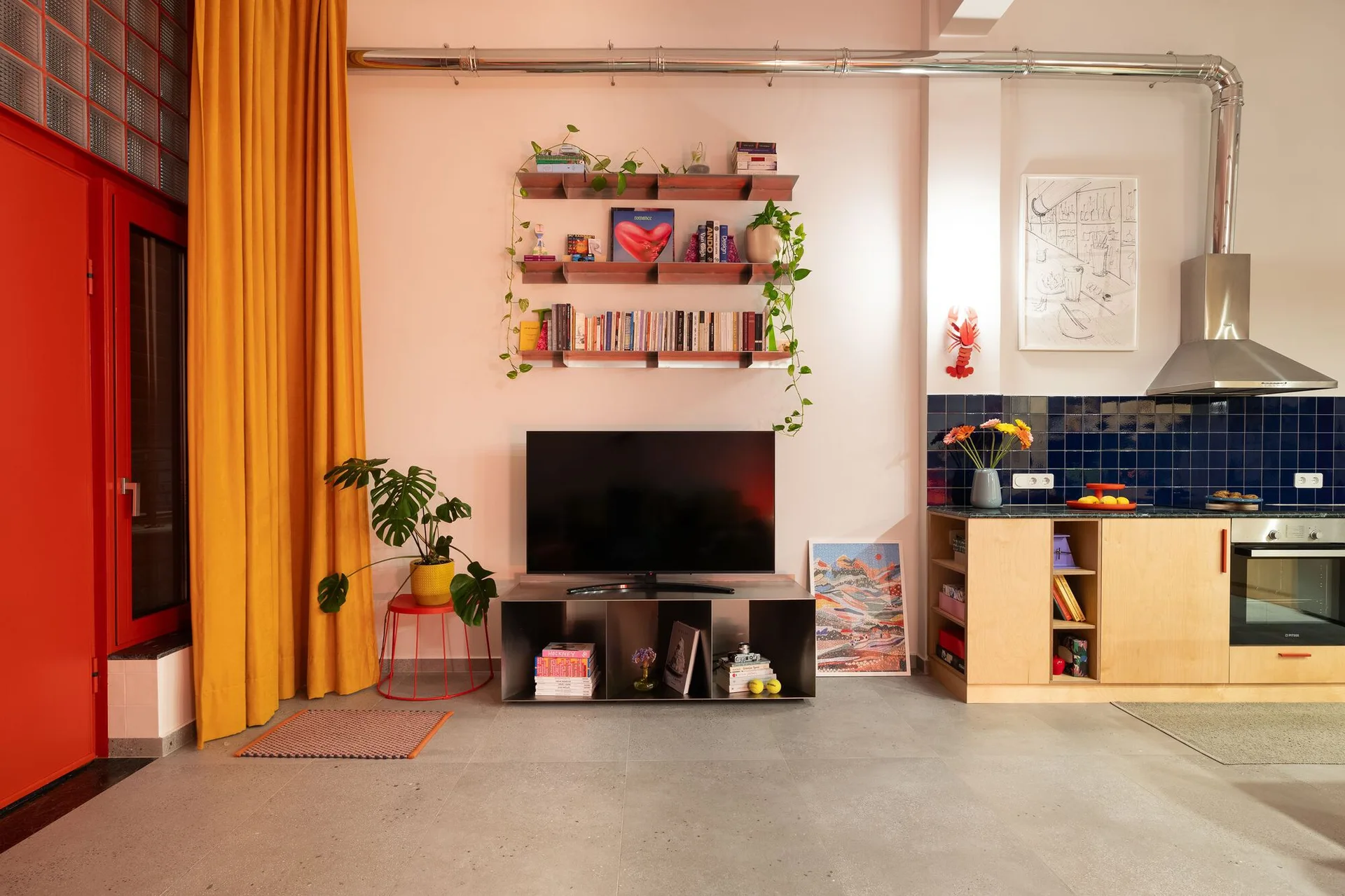 An open kitchen and living space with a television under metal wall shelves, a bright yellow curtain, and blue tiled kitchen backsplash.
